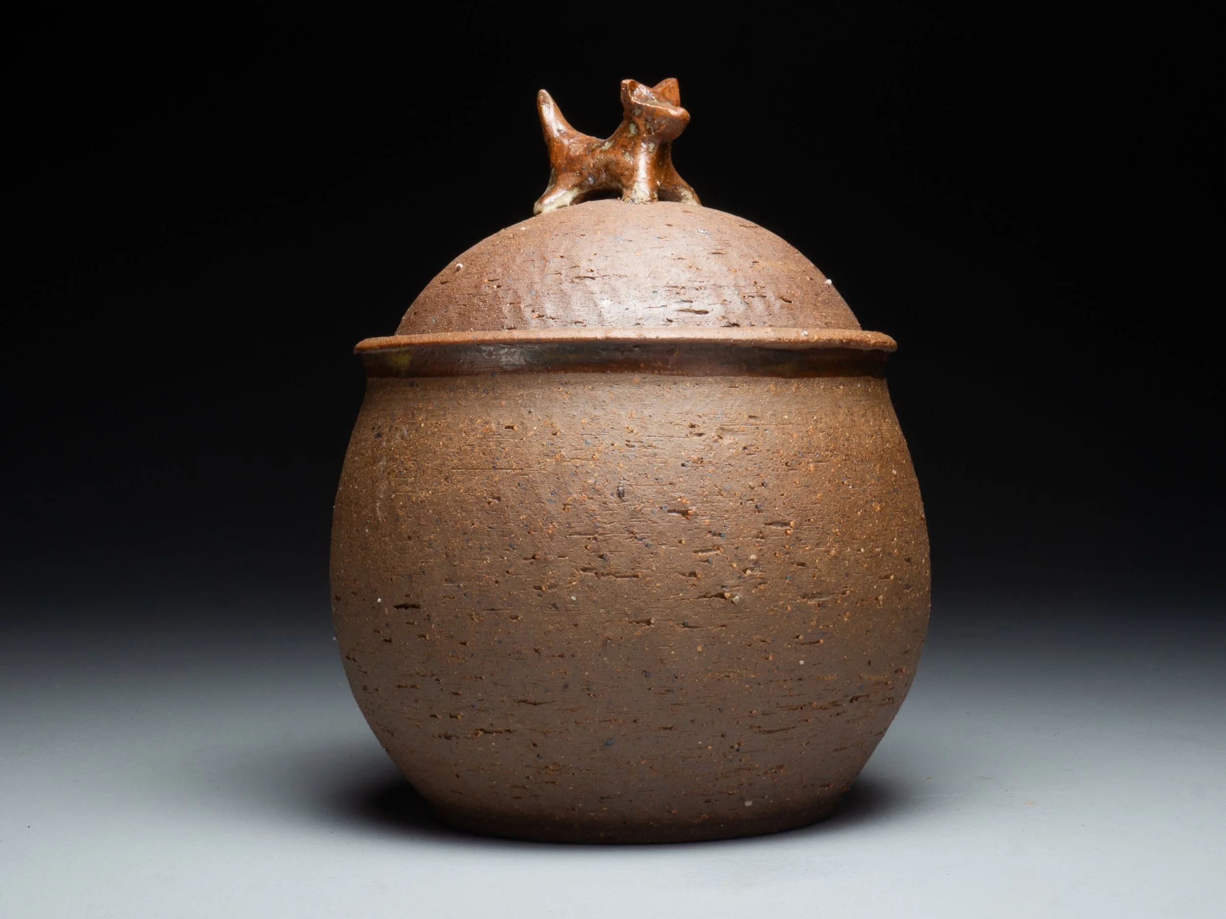 A rounded, reddish-brown ceramic vessel with a small dog figure on its lid, against a dark background.