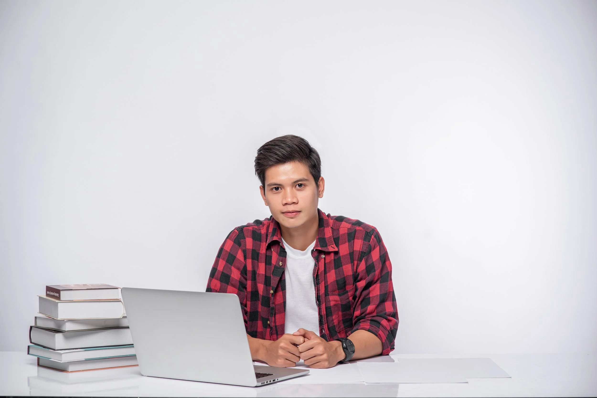 A young man with short dark hair wearing a red and black checkered shirt sitting at a white desk in front of a laptop, with a stack of books on his left, against a plain white wall.