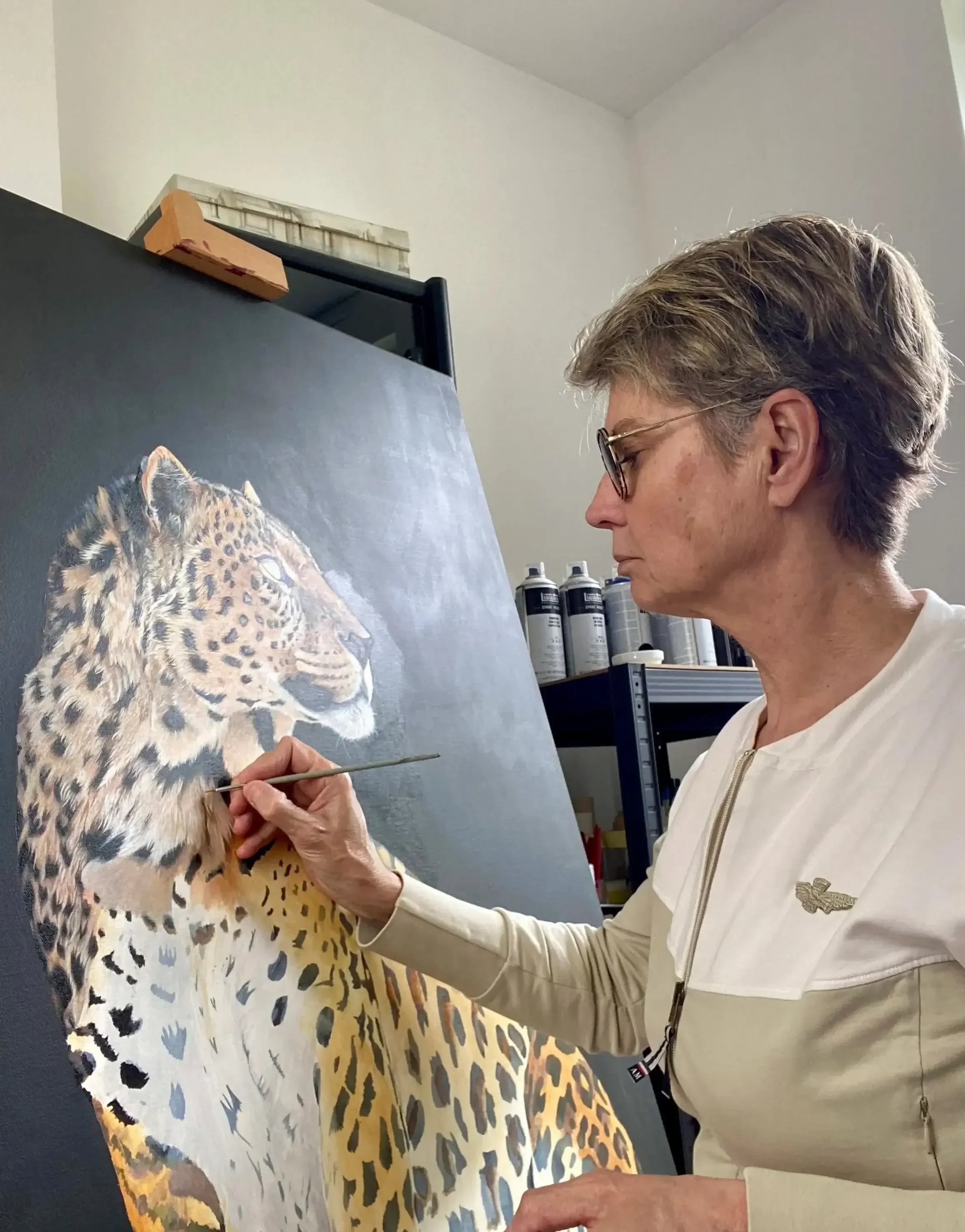 Lydia Mériaux, artiste peintre debout en train de peindre le portrait animalier à l'acrylique d'un léopard. Elle tient un pinceau. Elle est dans son atelier situé en Picardie.
