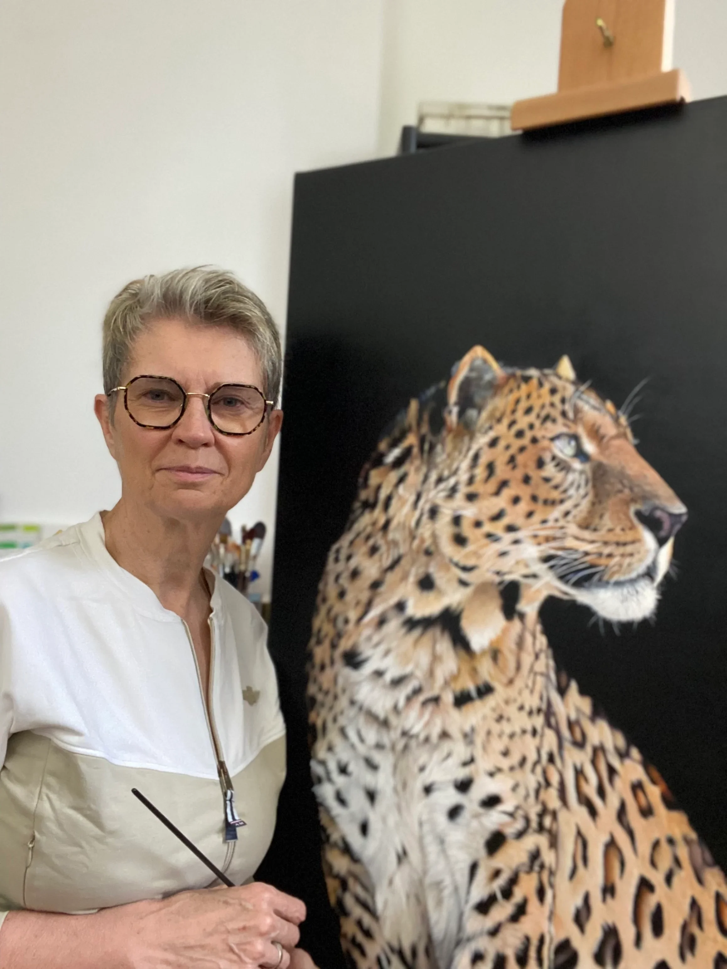 Lydia Mériaux, artiste peintre debout devant sa toile terminée intitulée "léopard beauté sauvage"". Elle tient un pinceau et regarde le spectateur en souriant. Elle est dans son atelier en Picardie.