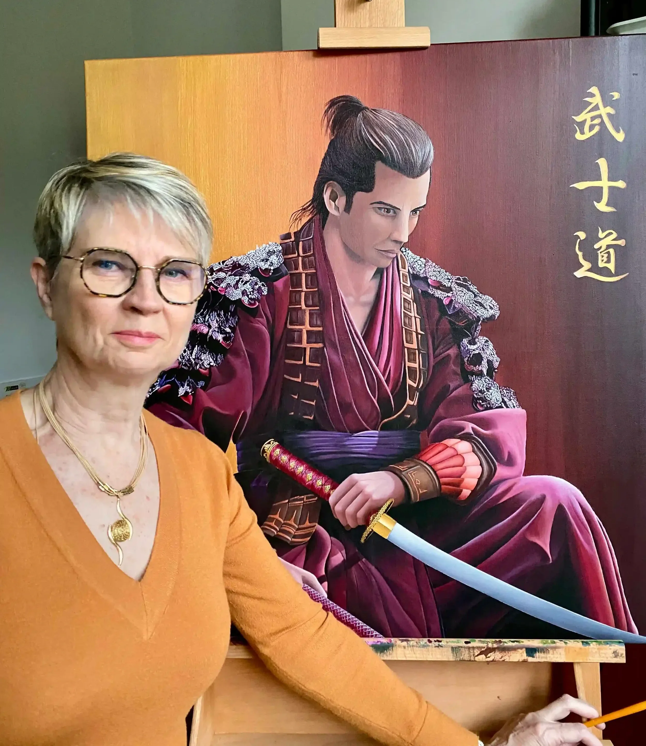 Lydia Mériaux, artiste peintre, portant un pull orange, tenant un pinceau, pose devant un chevalet sur lequel est posé sa toile représentant un samouraï en costume traditionnel.