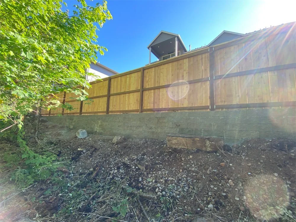 Residential backyard with new wooden fence on a sloped dirt and gravel terrain, bright sunny day with blue sky and sunlight glare.