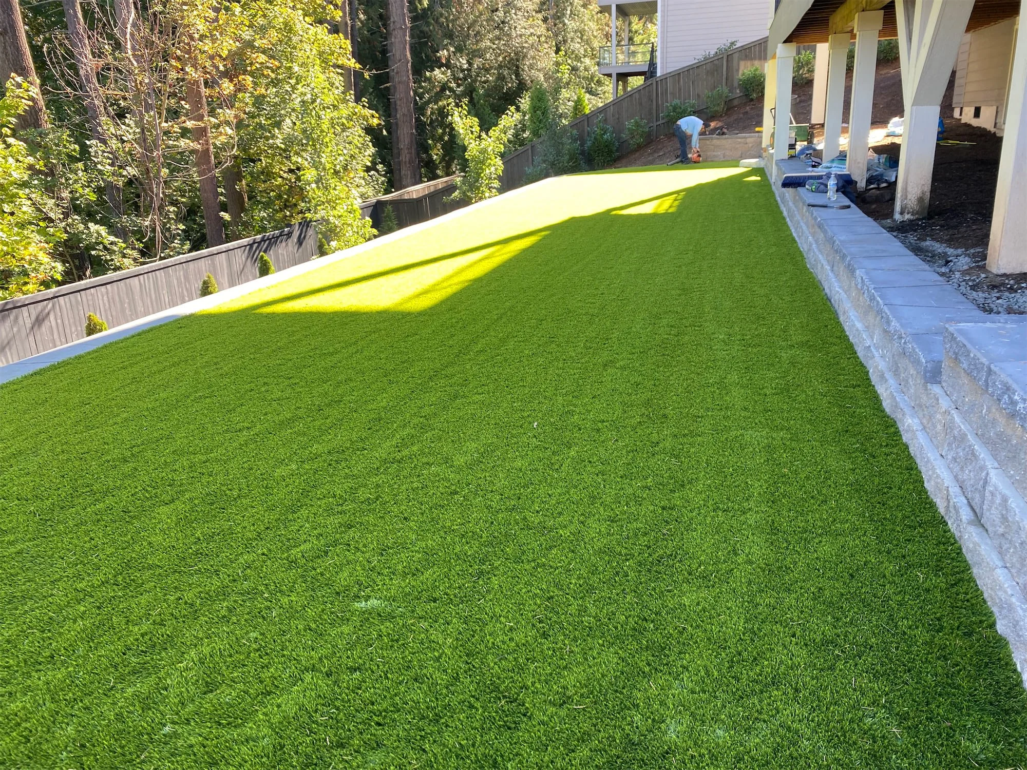 A backyard yard with freshly laid artificial grass next to a house with a raised porch, with workers installing the yard, and trees in the background.