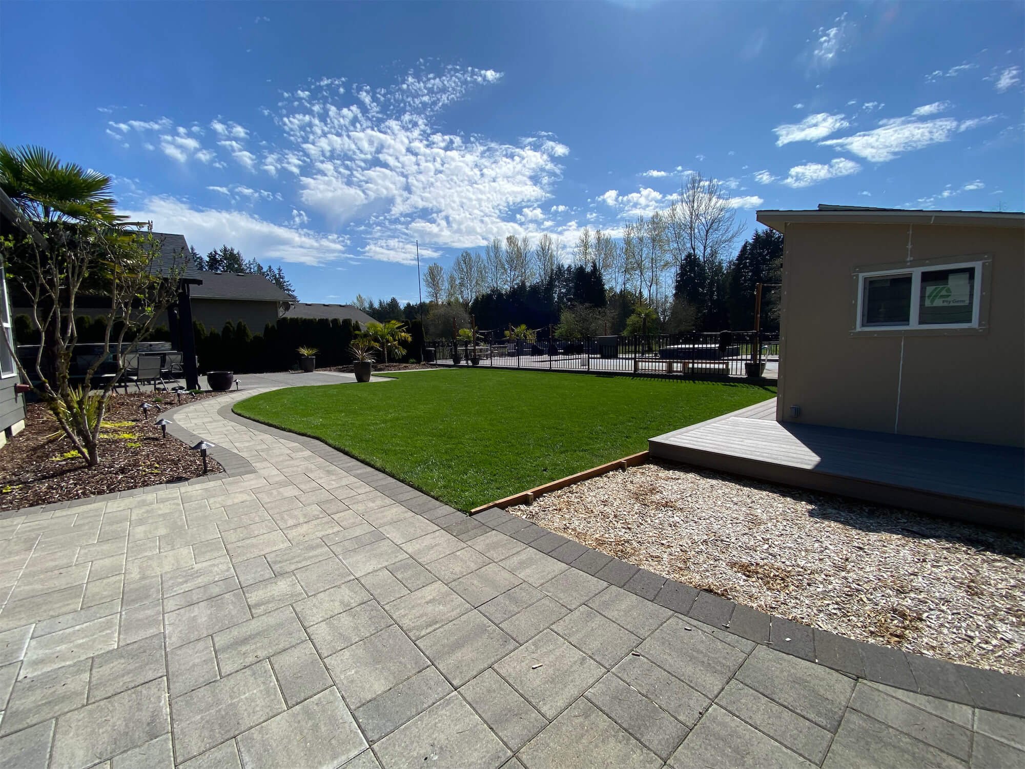 Backyard with a paved patio, a green lawn, potted plants, and a small shed. Mature trees with spring foliage in the background, partly cloudy sky.
