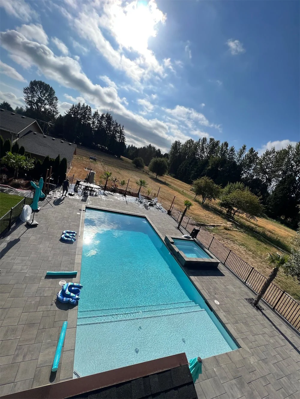 An empty outdoor swimming pool with a hot tub, pool floaties, and a pool cleaning tool on gray pavers, surrounded by trees, with a partly cloudy sky and sun overhead.