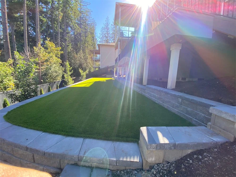 Backyard with artificial turf on a slope, stone edging, and a deck with columns, sunlight creating lens flare.