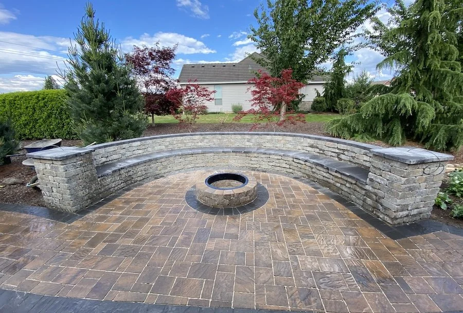 Curved brick bench surrounding a circular fire pit on a patio, with trees and a house in the background.