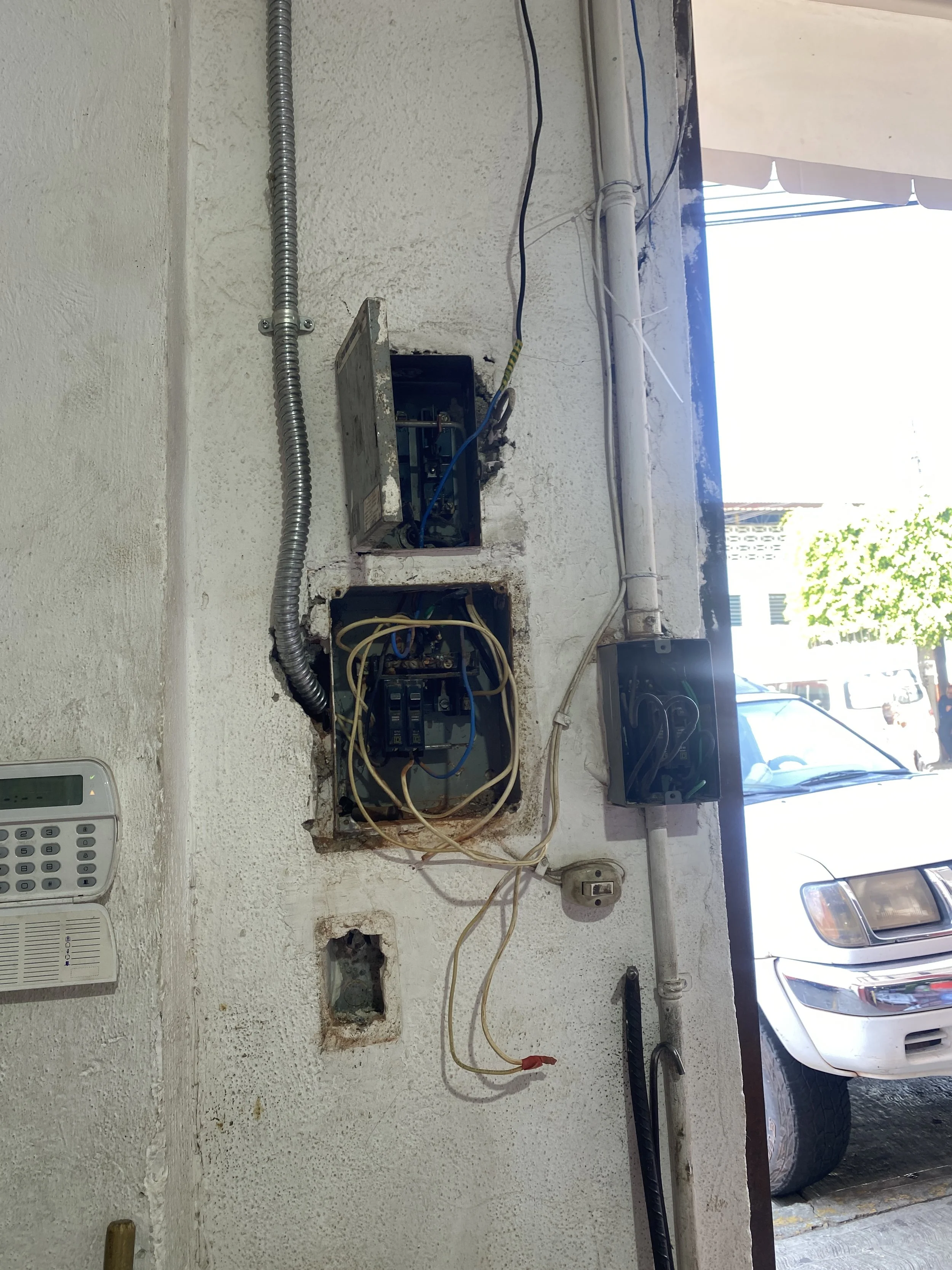 Trabajos de instalaciones eléctricas en una pared, con caja eléctrica, cables y ductos visibles, y una pared blanca de fondo, con una ventana que deja pasar luz.