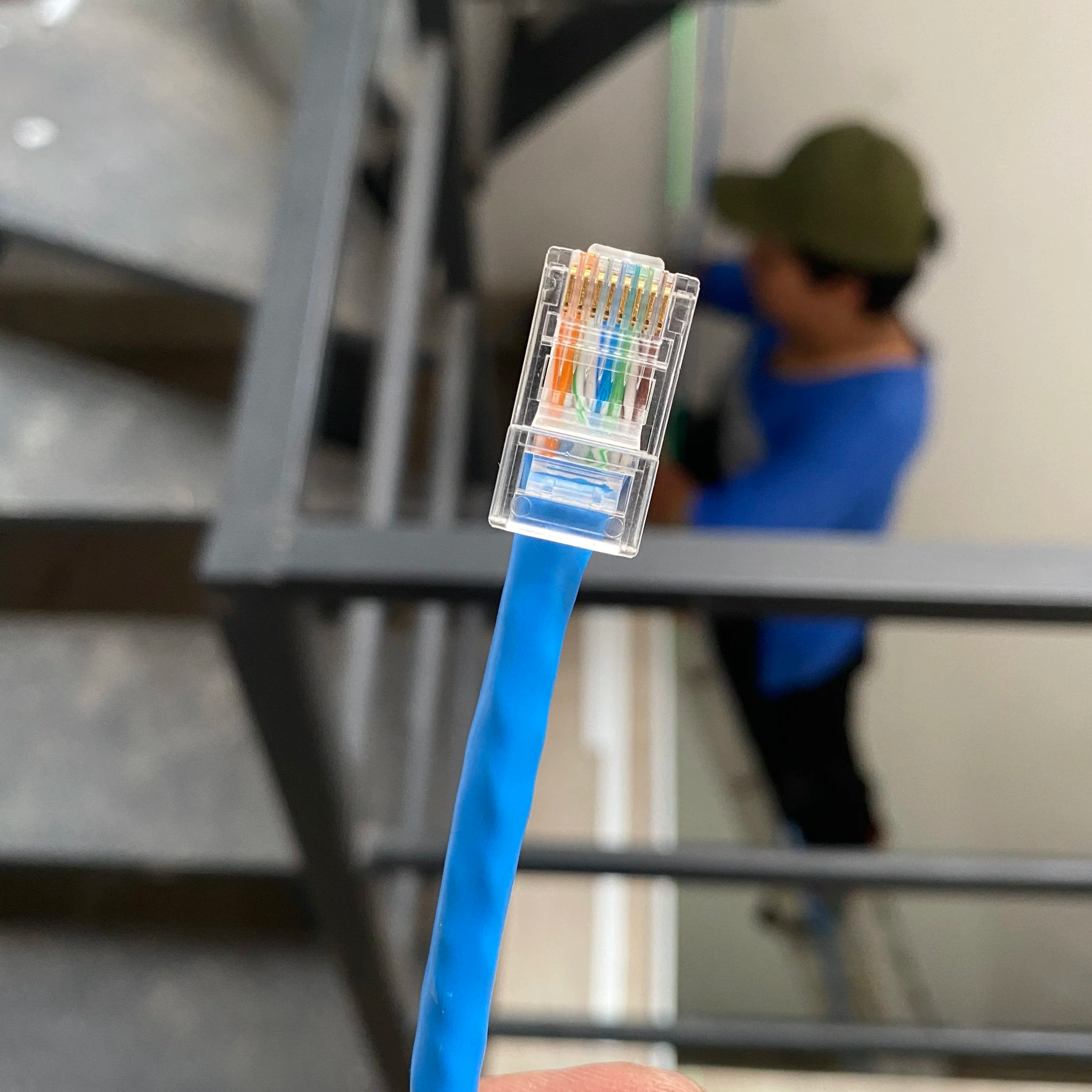 Un cable de red azul tiene un conector con varios cables de diferentes colores en una extensión de plástico transparente y está siendo sostenido por una mano. En el fondo, hay una estructura metálica y una persona con gorra y camiseta azul, parcialmente visible.