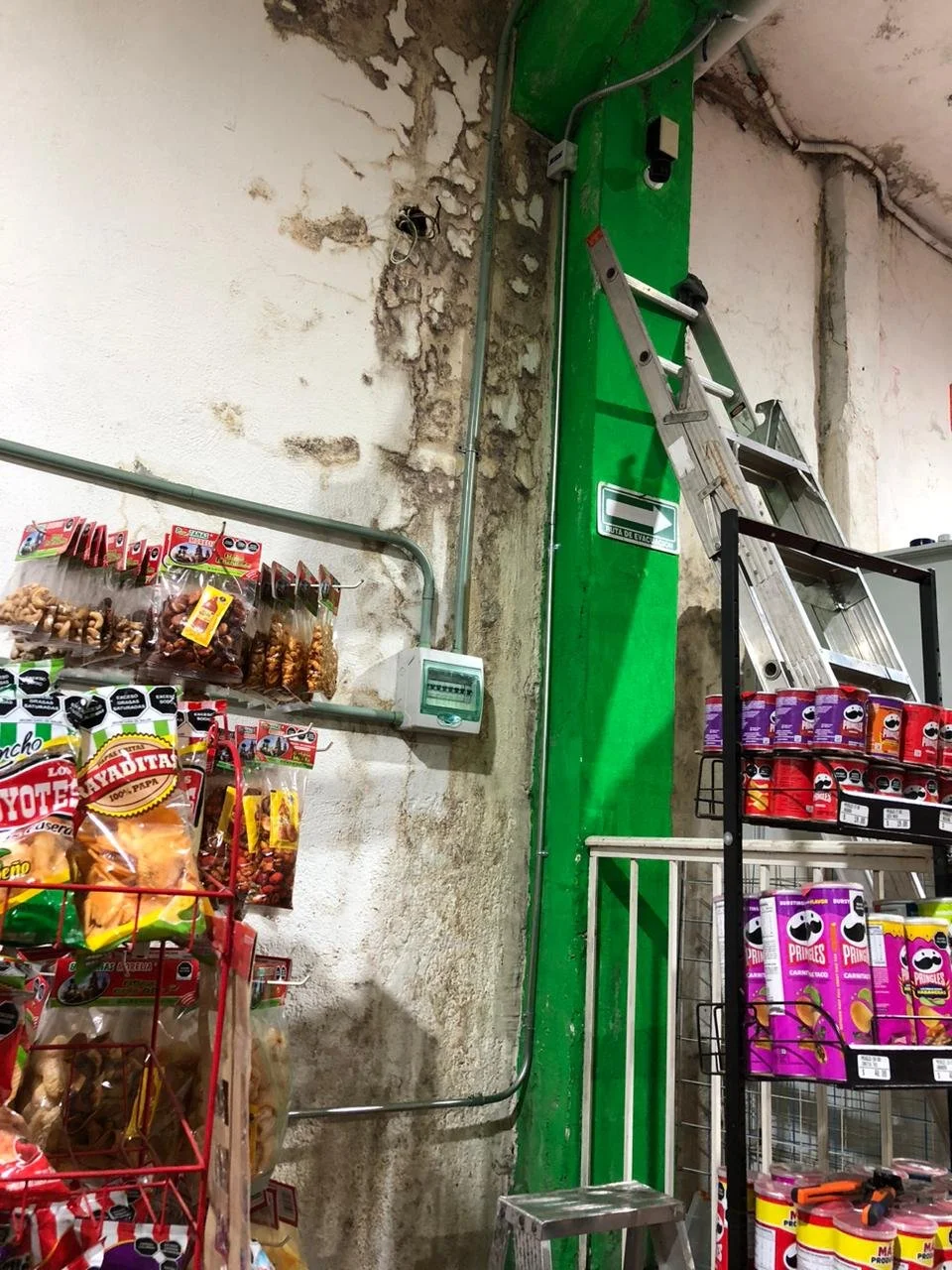 Estanterías con snacks y bebidas en la tienda, cerca de una escalera y una caja de fusibles, en un fondo de pared con desgaste y cableado aparente.