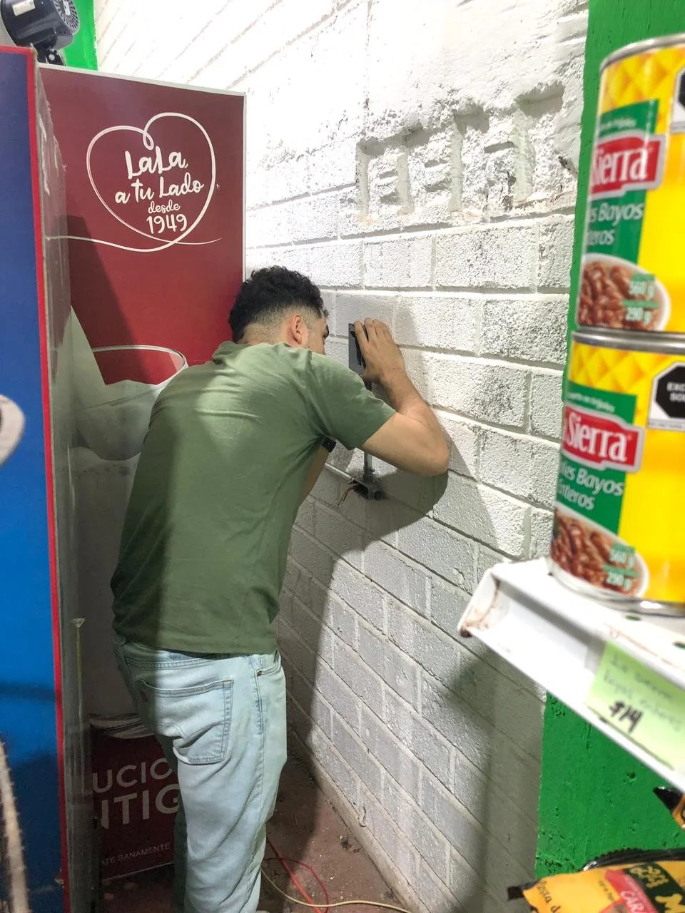 Un hombre trabajando en una pared de ladrillos blancos en una tienda, con frascos de alimentos en primer plano y un cartel rojo con texto en español en el fondo.