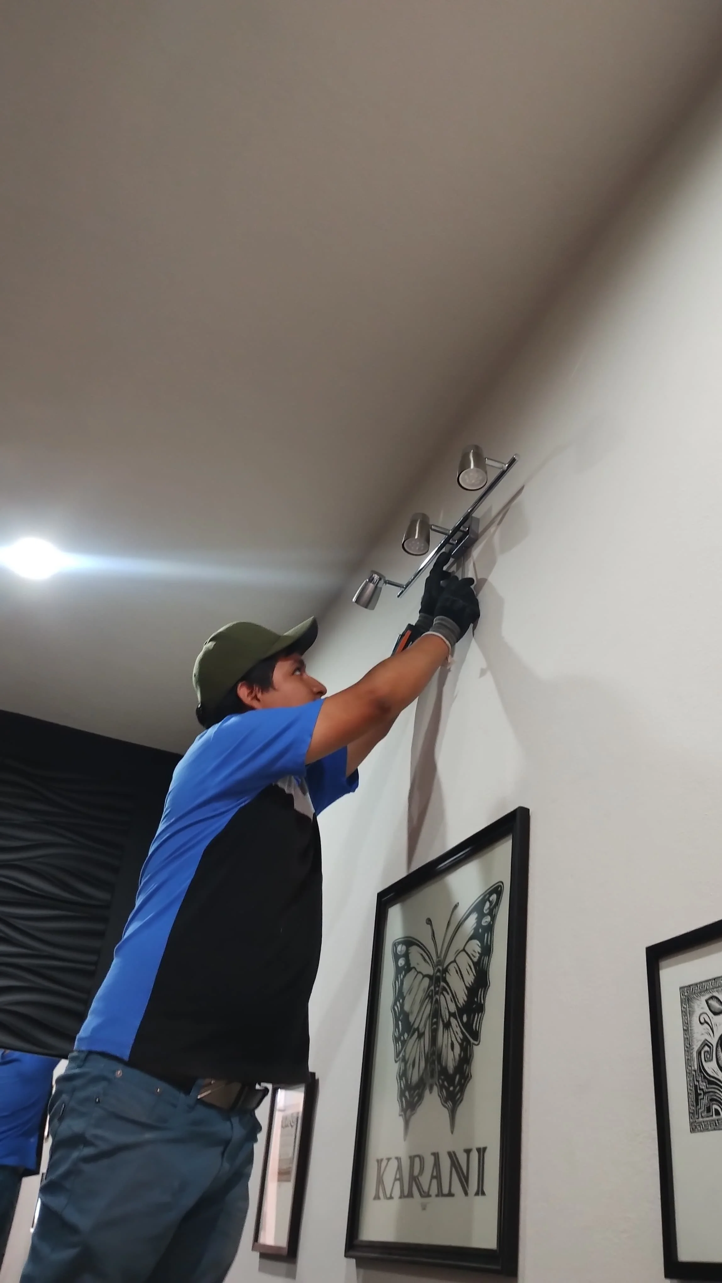 Joven instalando una lámpara en la pared interior, con cuadros decorativos y un cartel que dice "KARANI" en la pared.