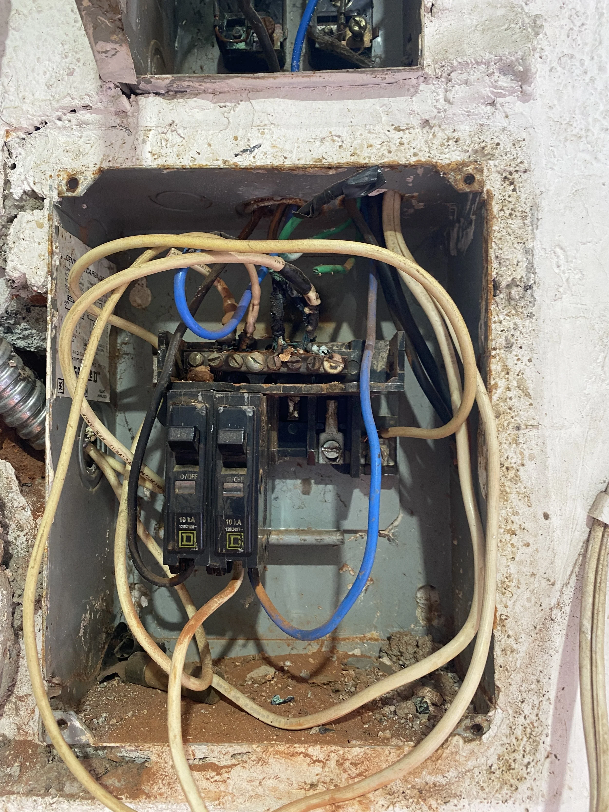 Caja eléctrica con cables desgastados y conexiones oxidadas, empotrada en una pared de yeso con daños y polvo en el suelo.