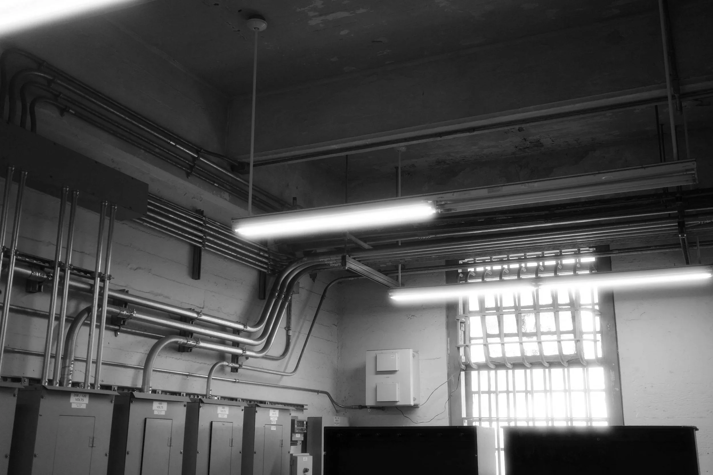 Imagen en blanco y negro de un cuarto de instalaciones eléctricas, con varias tuberías metálicas, un panel de control y ventanas que dejan pasar la luz natural.