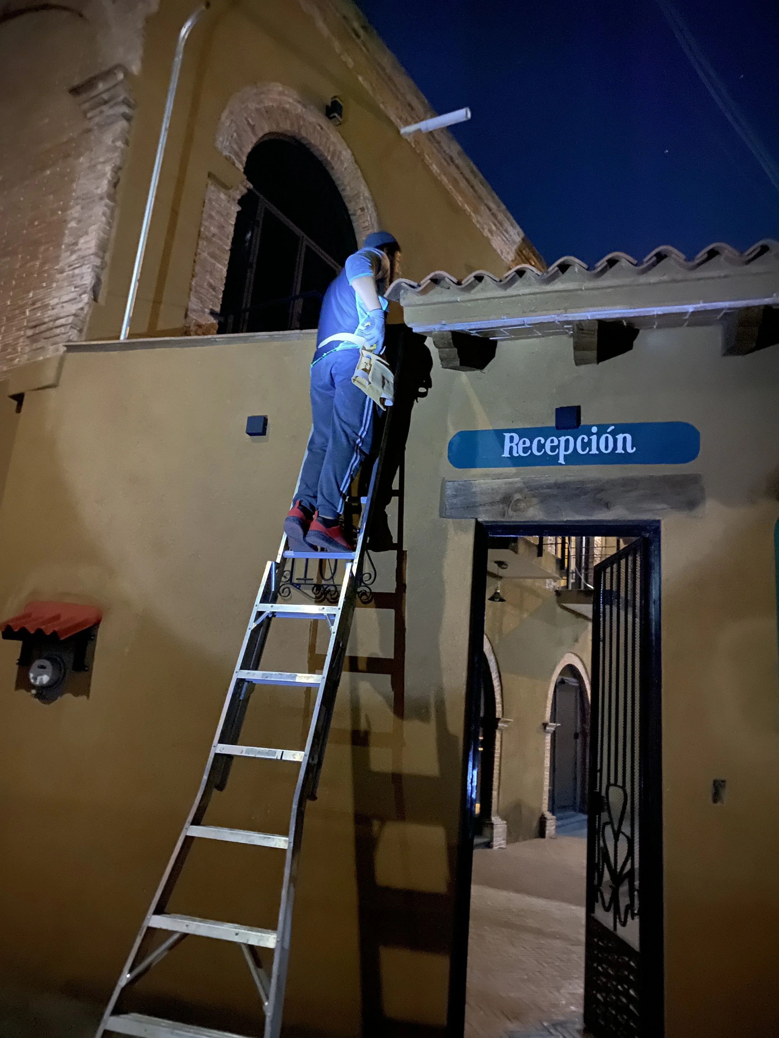Persona en una escalera subiendo a la entrada de un edificio con un letrero azul que dice "Recepción". Es de noche y hay una luz brillante alumbrando la escena.