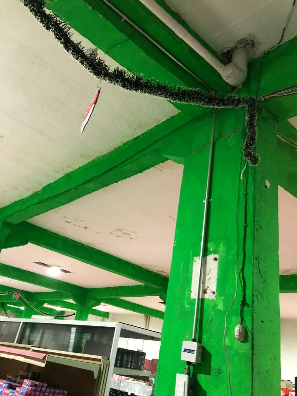 Interior de un local con estructura de columnas y techos pintados de verde. Se observa un canaleta con cableado, una caja de conexiones y una manguera negra enroscada en la esquina del techo.