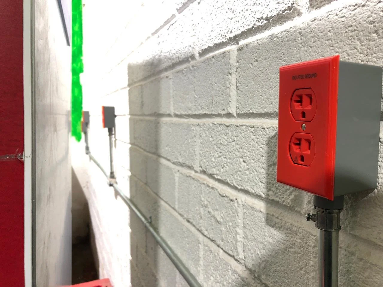 En una pared de ladrillos blancos, hay un enchufe rojo de emergencia con dos tomas eléctricas y un riel de cables. También se puede ver una luz verde en el lado izquierdo.