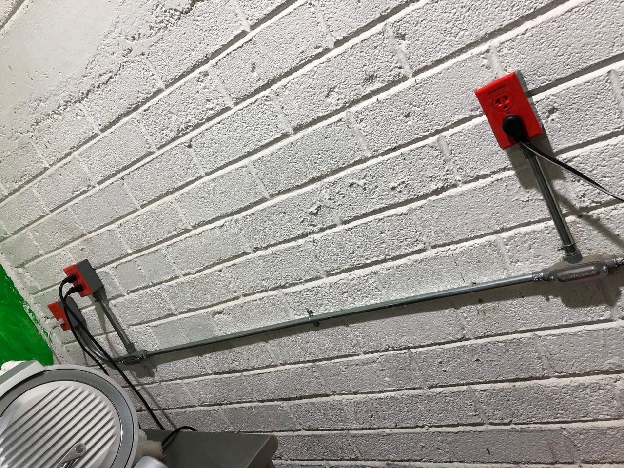 Instalaciones eléctricas en pared de ladrillo pintado de blanco, con conductos y enchufes rojos de protección contra pérdidas, y una máquina de ejercicio parcialmente visible en la esquina inferior izquierda.