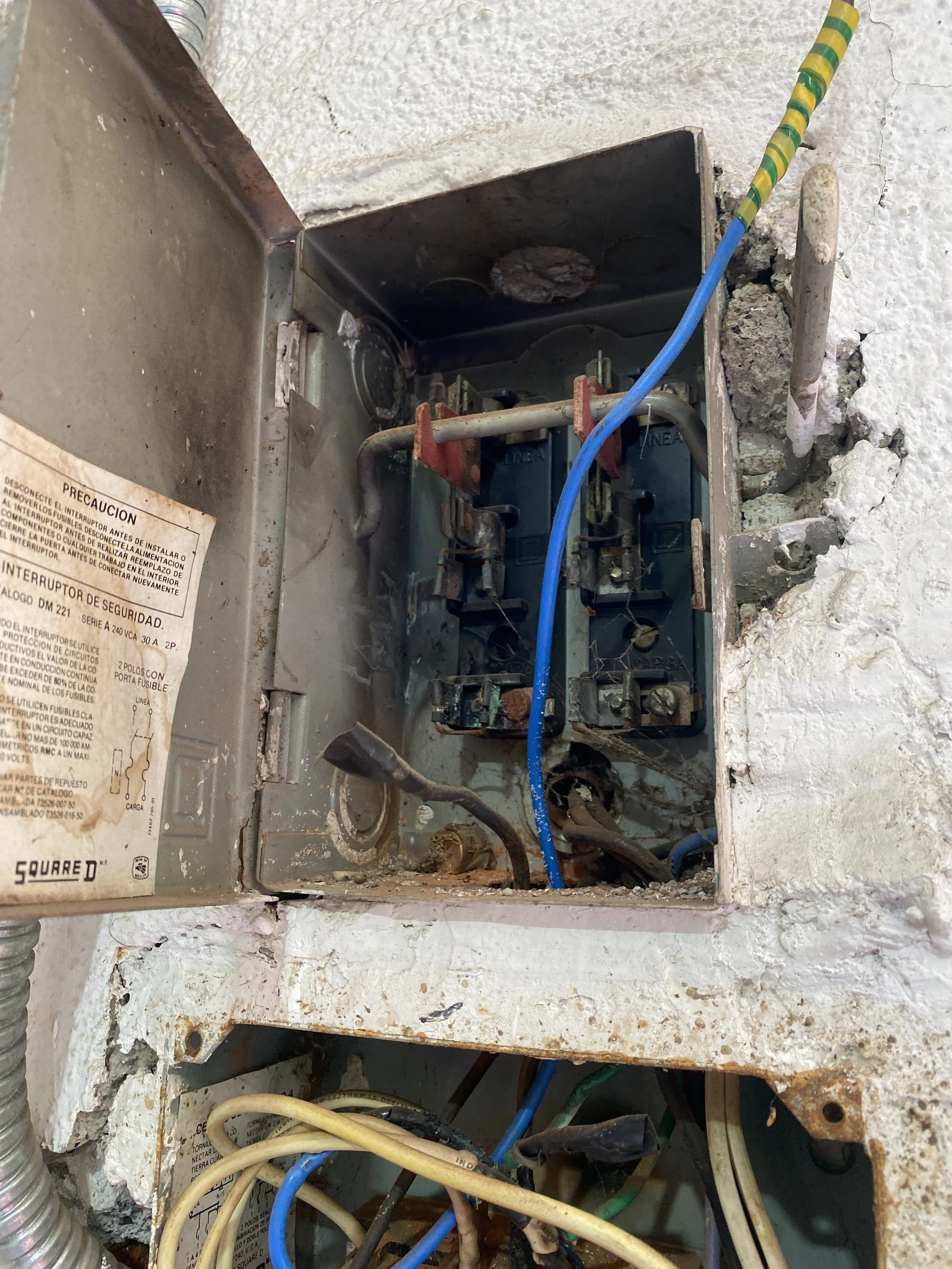 Caja de interruptor eléctrico con cables conectados y enchufes en una pared de yeso dañada con grietas y restos de cemento.