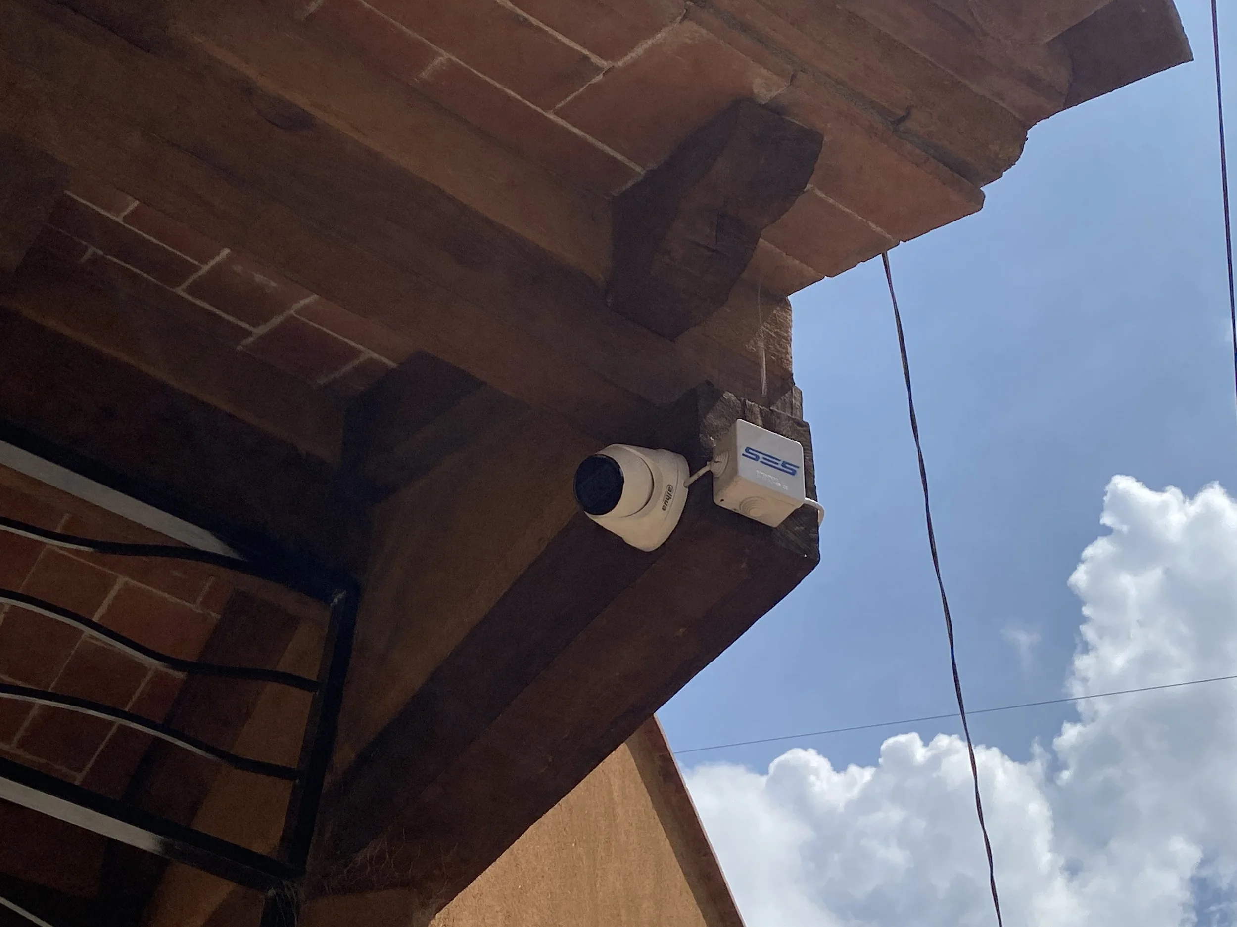 Foto del cielo azul con algunas nubes visibles, junto a un edificio de ladrillos con una cámara de seguridad instalada en la esquina y cables eléctricos colgando.