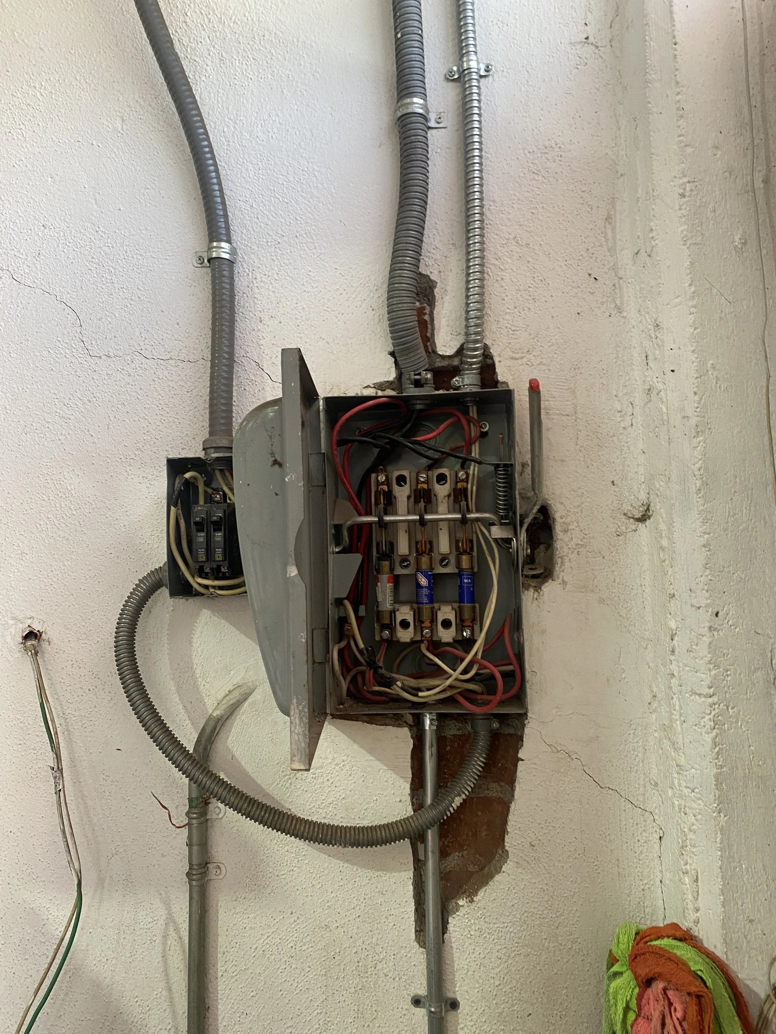 Cuadro eléctrico abierto con cables y componentes internos en la pared, y cables en el suelo a la izquierda, con un paño de color naranja, verde y rosa en la esquina inferior derecha.