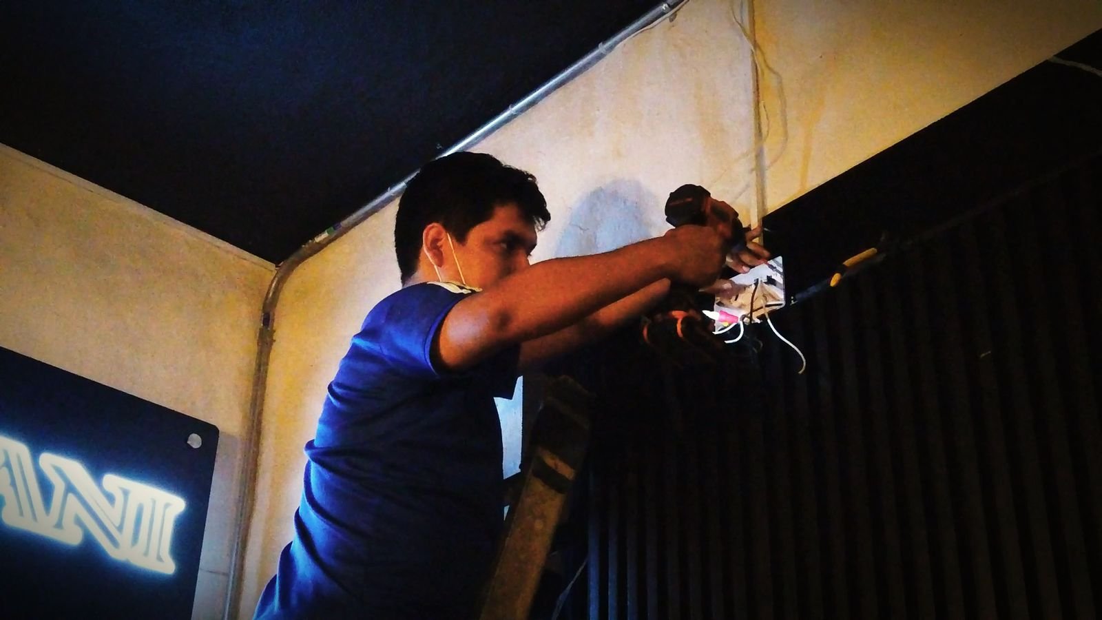 Un técnico instalando o reparando cables en una pared, usando herramientas y de espaldas, en un entorno con paredes de tonos claros y un letrero luminoso en la parte inferior izquierda.