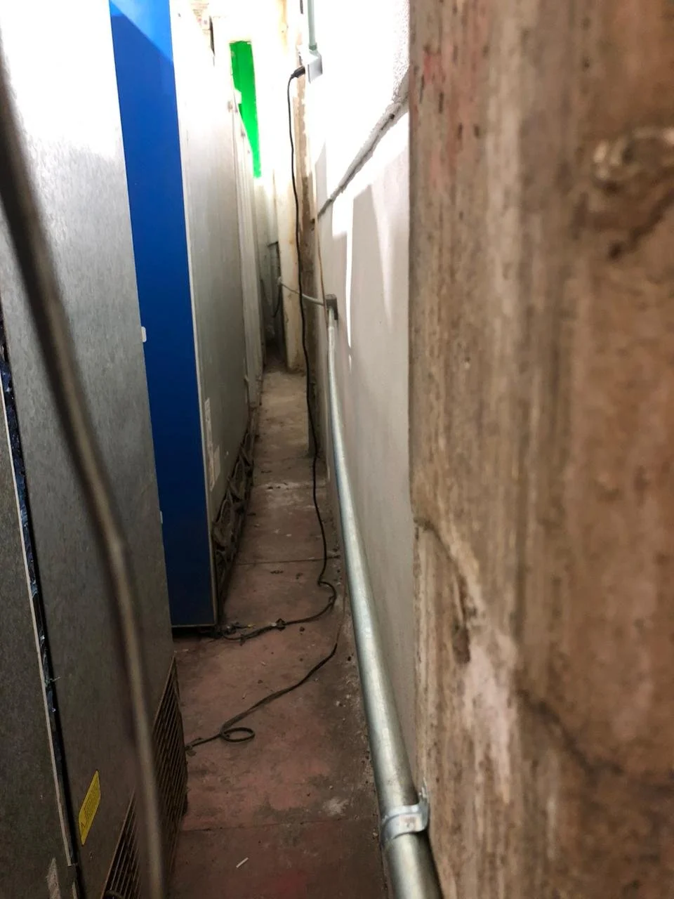 Pasillo estrecho entre paredes de ladrillo y objetos eléctricos, con cables visibles en el suelo.