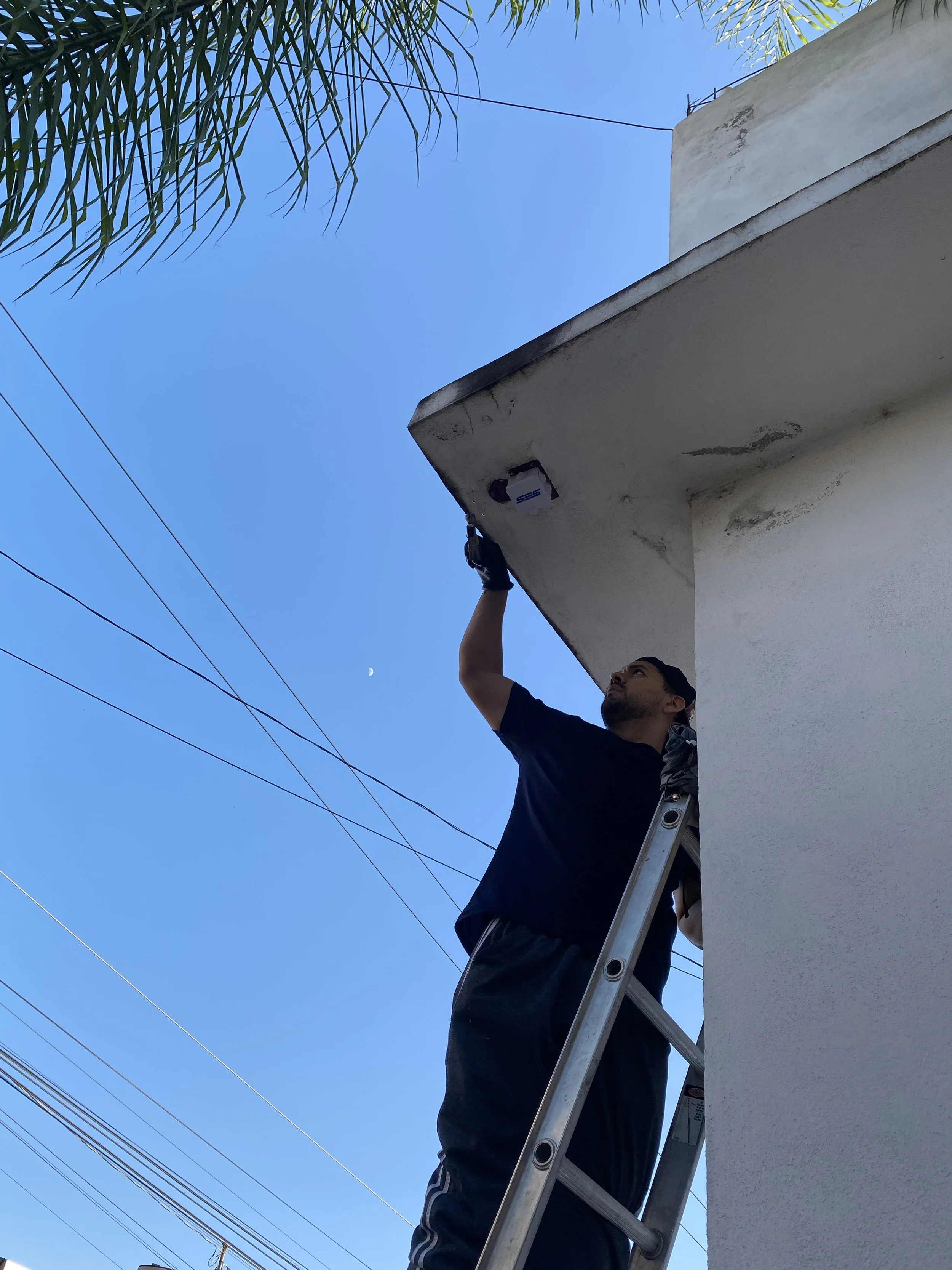 Hombre en una escalera realizando mantenimiento en la parte superior de un edificio, con cables eléctricos y un cielo claro de fondo.