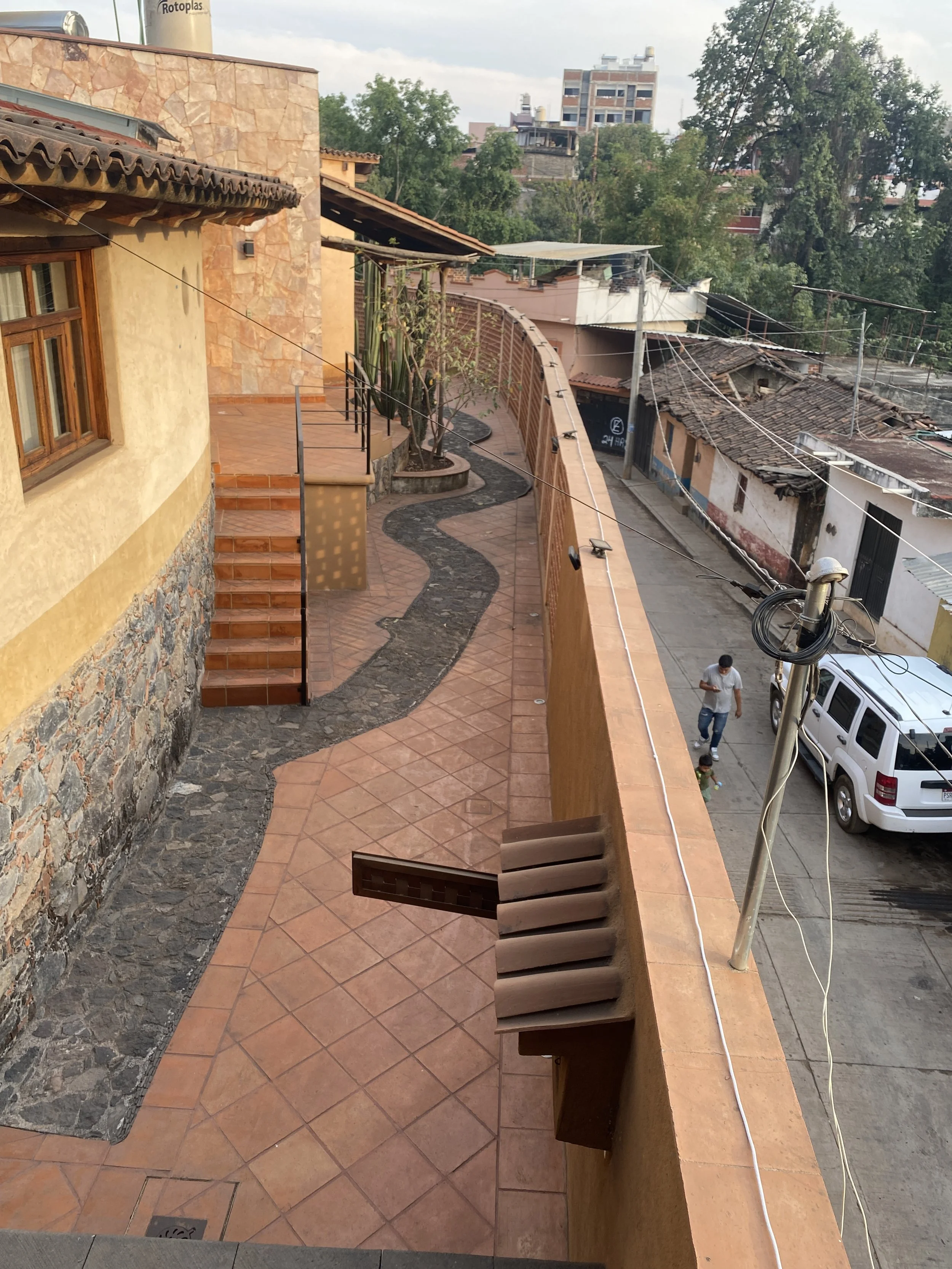 Área exterior de una casa con pasillo de baldosas de terracota, vallas de ladrillo y escaleras, con árboles y edificios en el fondo.