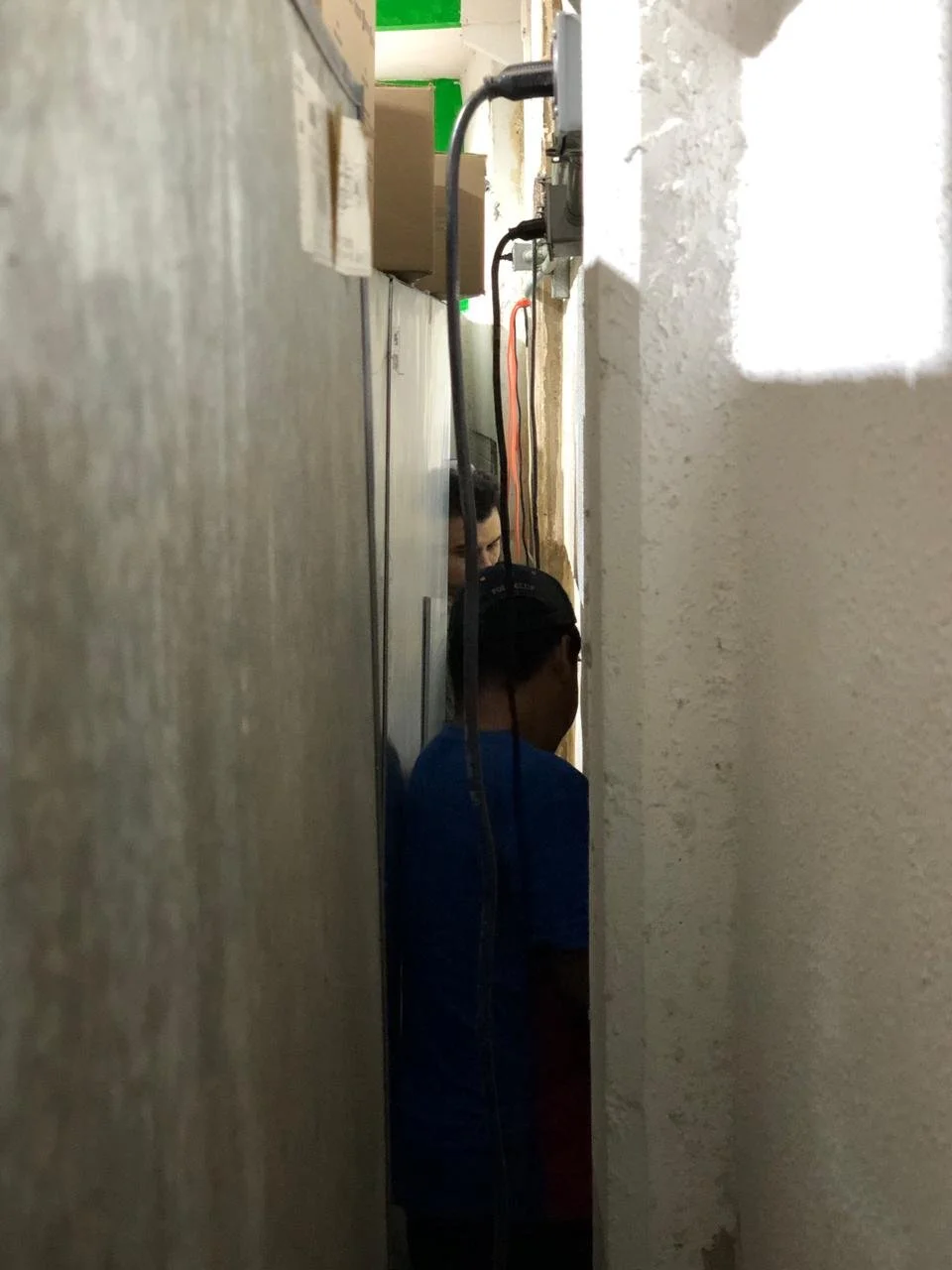 Tres personas en un espacio estrecho entre paredes, con cables y enchufes visibles en la pared, una de ellas usando un casco y otra mirando hacia abajo.