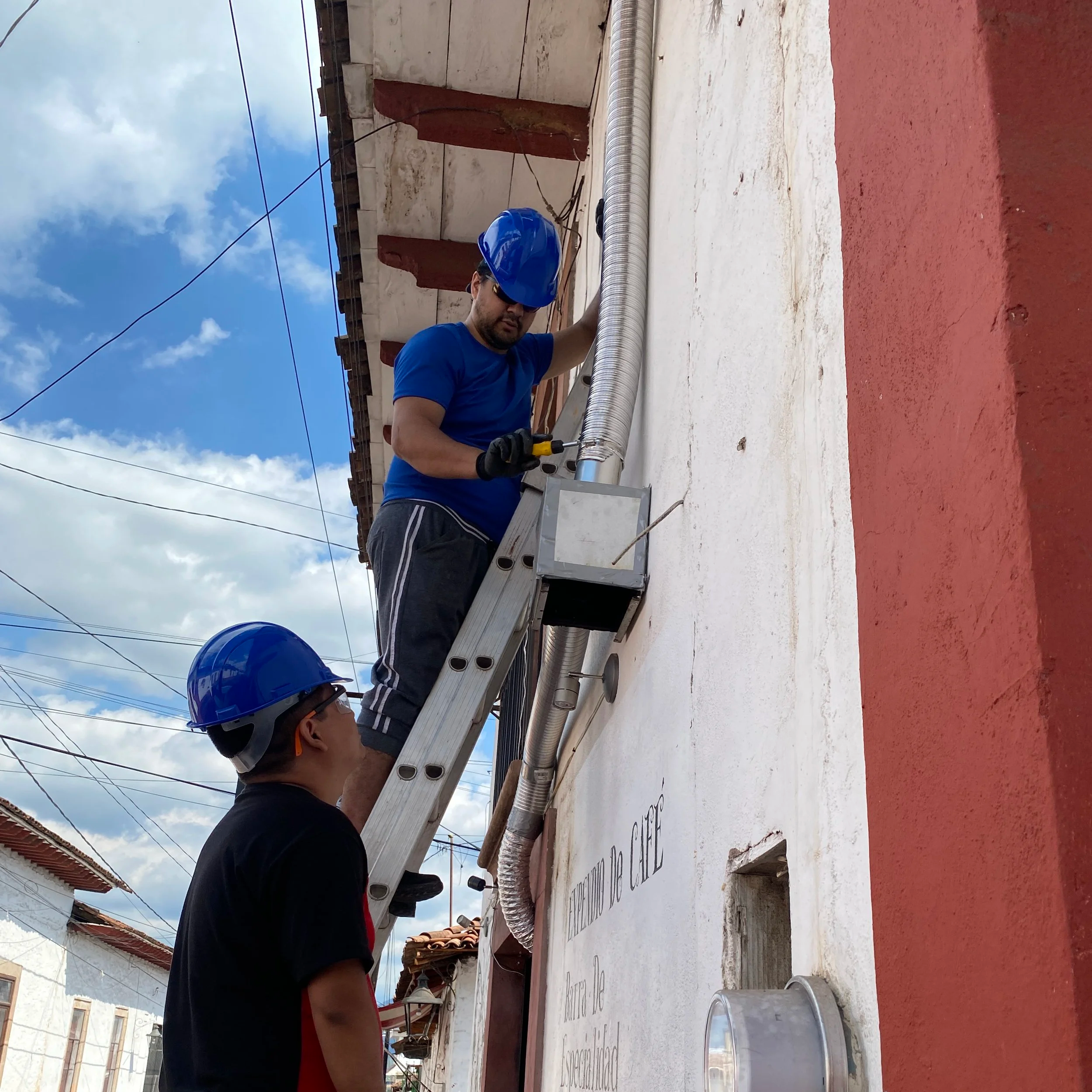 Varón con casco azul y gafas instalando tubos de ventilación en una azotea con cielo parcialmente nublado.