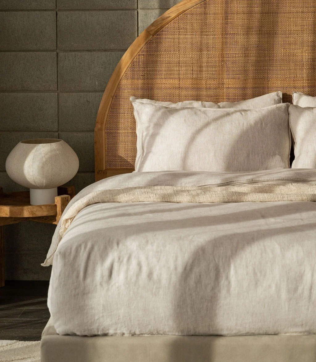 A bedroom with a bed featuring beige linens, a curved wooden headboard with rattan inset, a bedside table, and a modern white table lamp, against a dark gray stone wall.