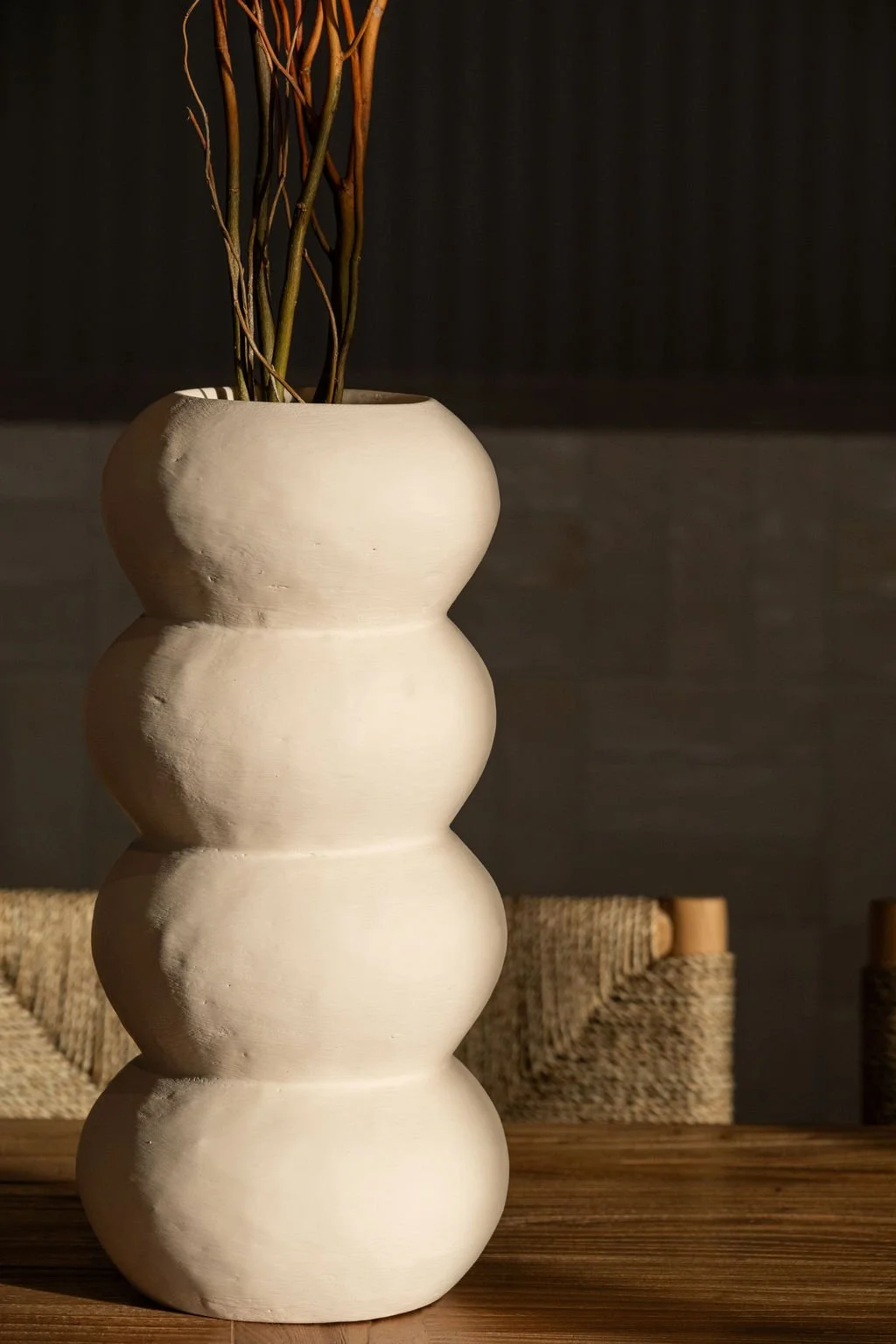 A tall, sculptural white ceramic vase with a rounded, stacked shape holding dried branches, placed on a wooden surface with a dark, textured background.
