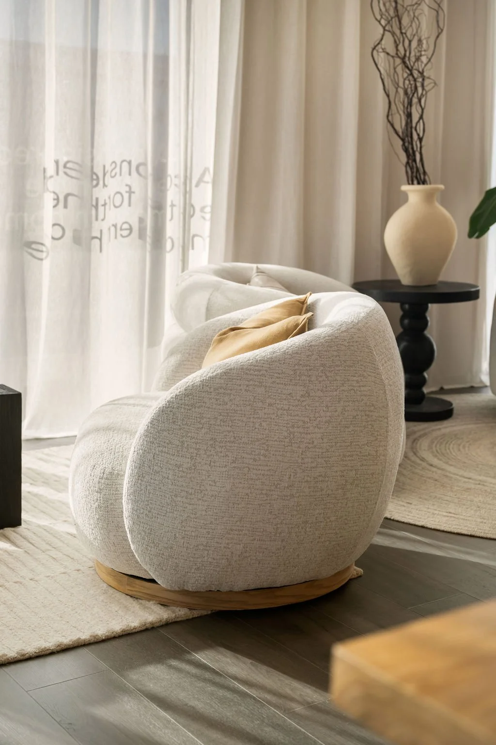 A cozy living room corner featuring a beige, textured armchair with tan cushions, a black round side table with a large beige vase filled with dried twigs, white curtains with faint text, and a circular woven rug on light-colored wooden flooring.