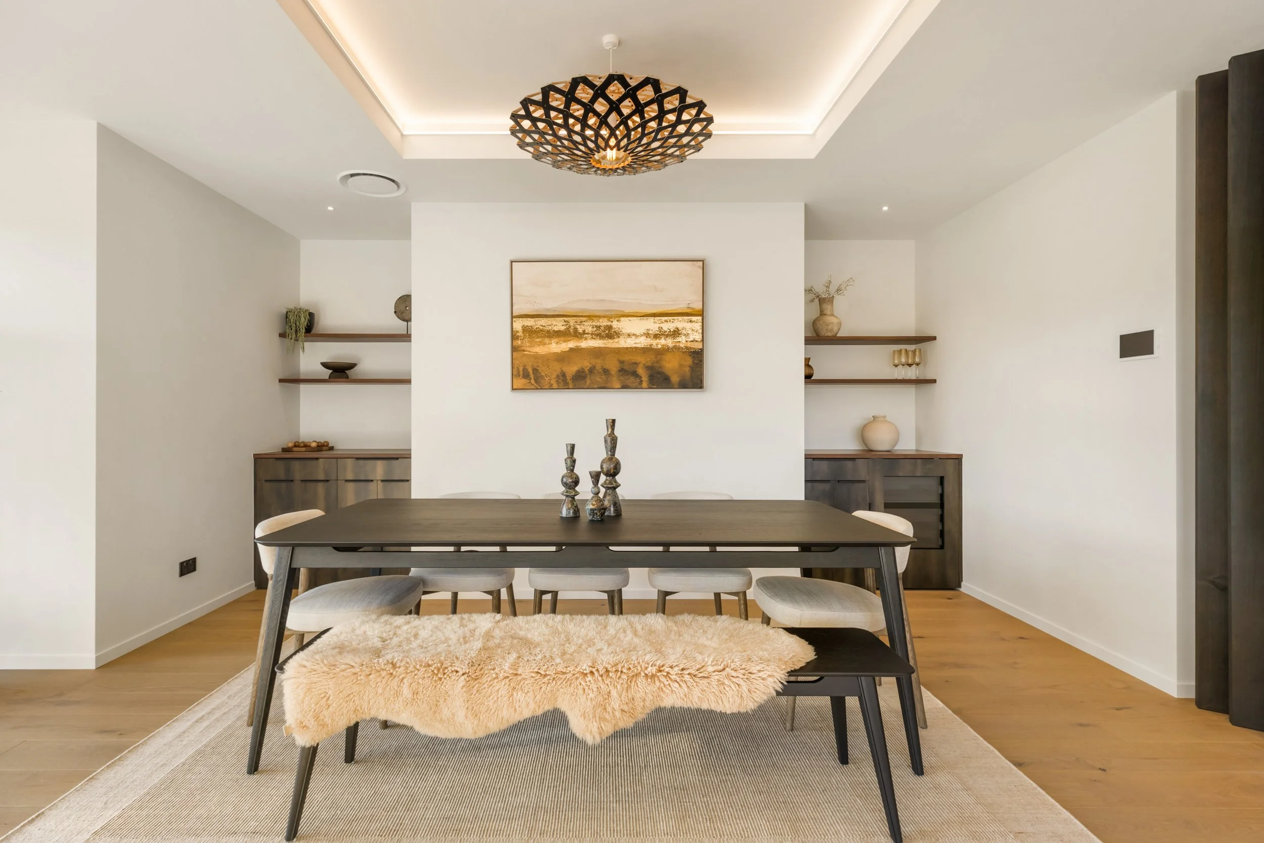 Modern dining room with a wooden table, six upholstered chairs, a fluffy beige rug, wall-mounted shelves with decorative items, and a painting on the wall, illuminated by a statement chandelier.