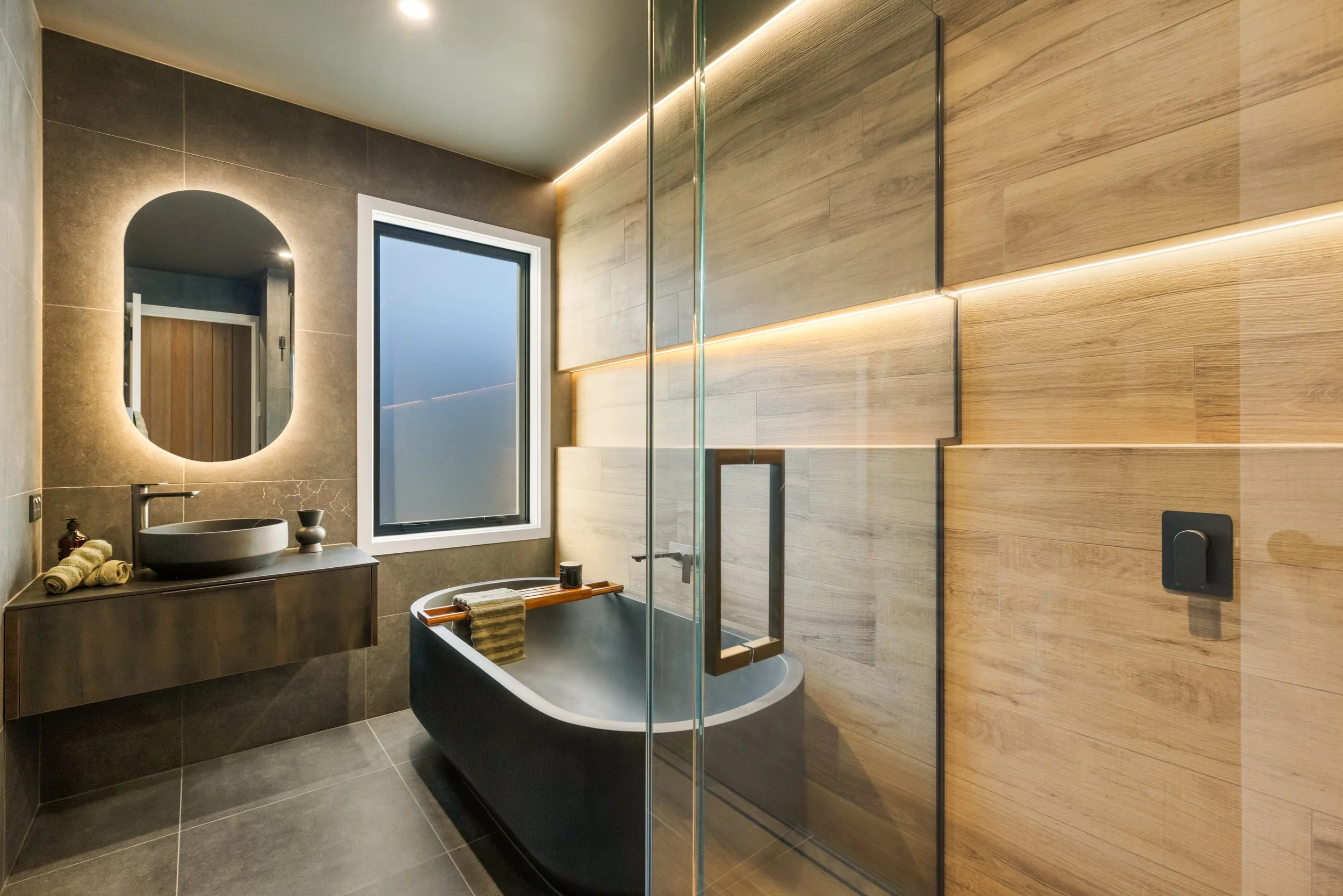 Modern bathroom with a black clawfoot tub, glass shower enclosure, wooden wall panels, and a gray stone vanity with a vessel sink and mirror with backlighting.