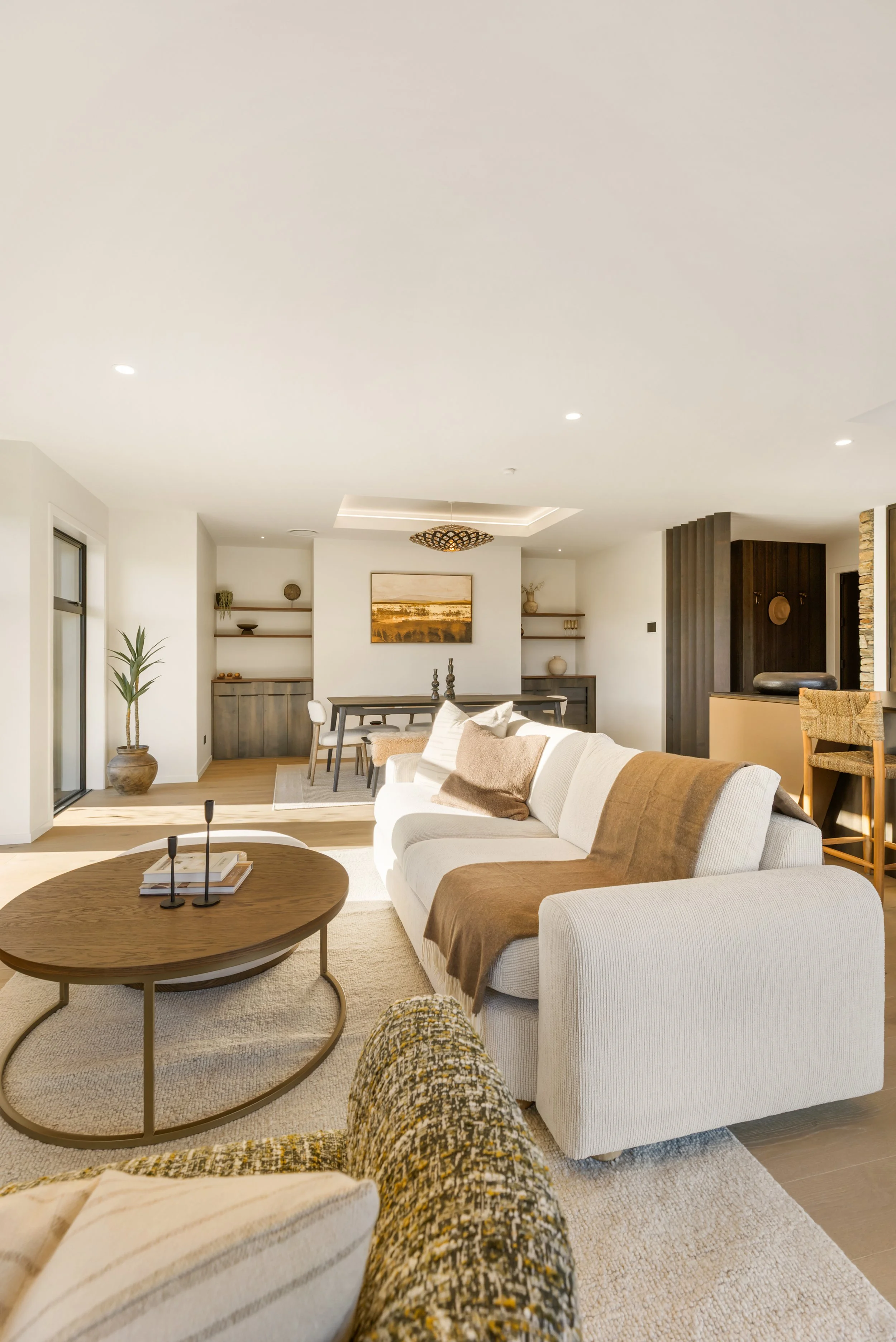 Modern living room with white couches, a round wooden coffee table, and open books on it. There's a beige carpet, a patterned pillow, and a person’s legs wearing colorful pants in the foreground. The background features minimalist decor including wal