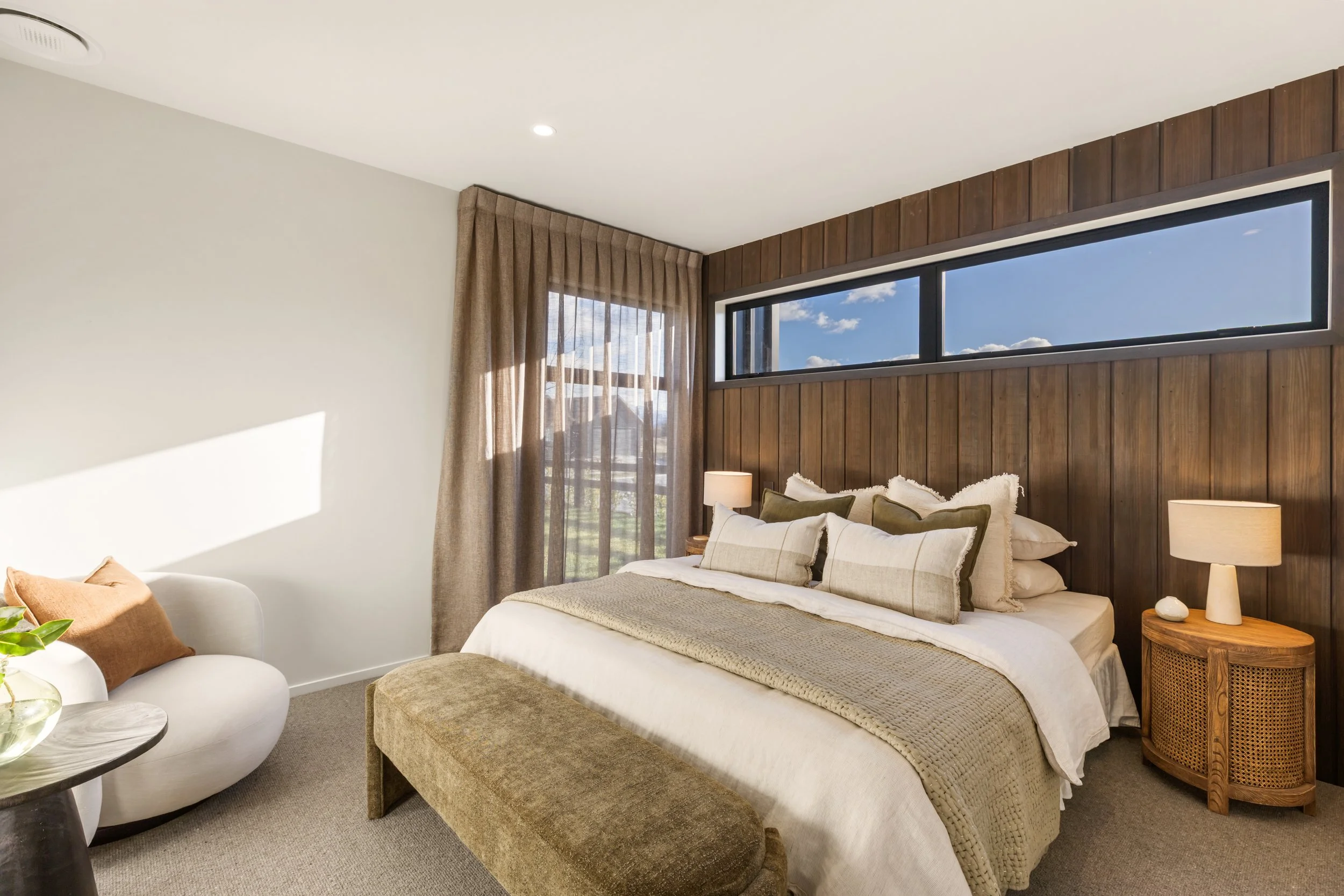 A bedroom with a bed, pillows, lamps, wooden wall paneling, large window with curtains, and a sky view.