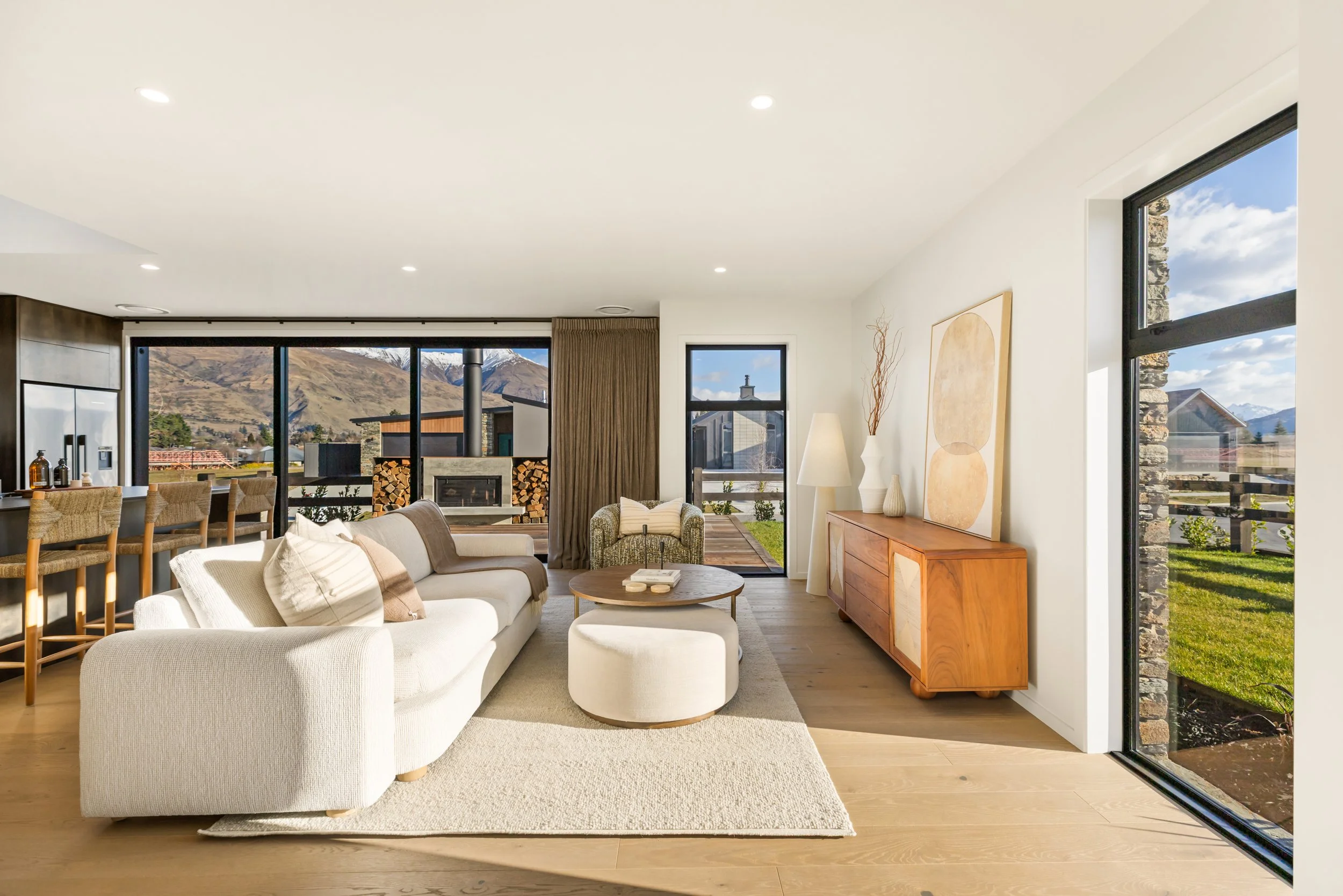 Bright living room with white sofa, round ottoman, wooden sideboard, large windows showing mountains, firewood stacked near fireplace, art on wall, and outdoor view of green yard and blue sky.