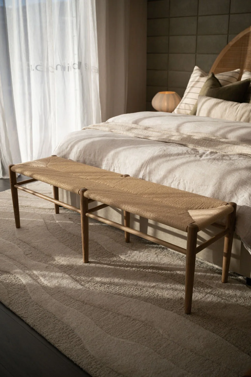 A bedroom with a bed, a wooden bench at the foot of the bed, a bedside table with a lamp, and sheer curtains.