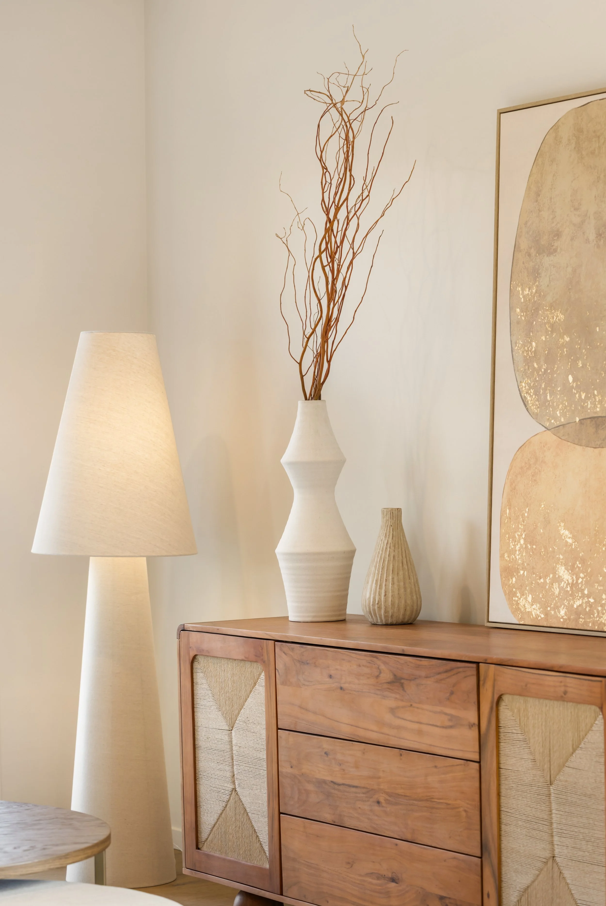 Decorative interior scene with a wooden sideboard, a beige table lamp, vases with dried branches, and abstract wall art in neutral tones.