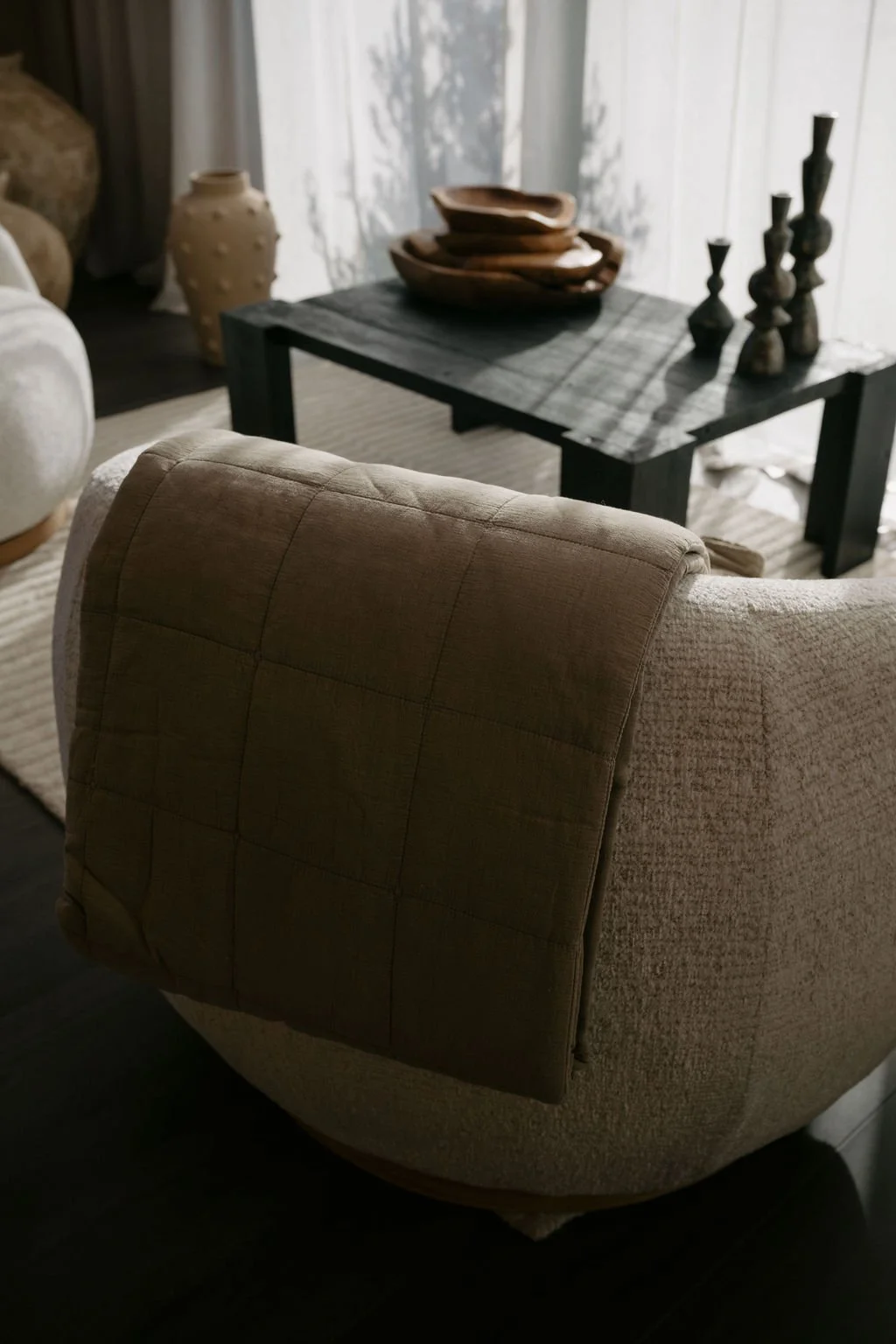 A beige upholstered armchair in the foreground of a living room with a dark wooden coffee table decorated with bowls and candlesticks.