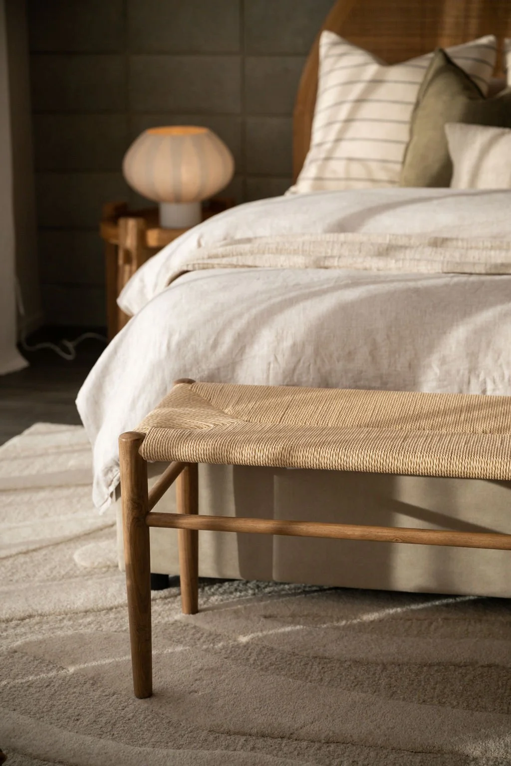 Close-up of a bedroom with a bed featuring white linen and striped pillows, a wooden bench at the foot of the bed, a rug, and a bedside table with a lamp in a warmly lit room.