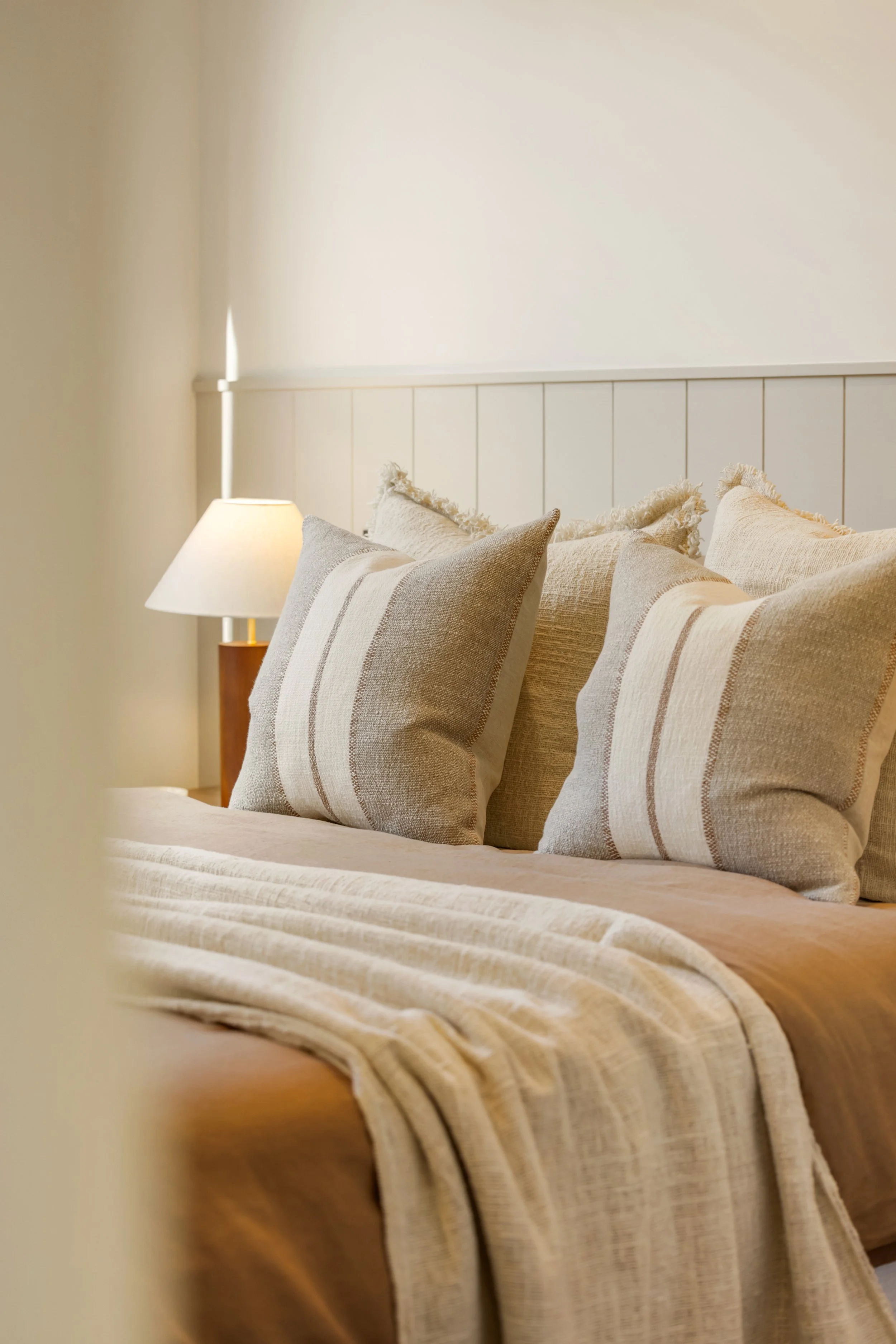 A neatly made bed with beige and cream striped pillows and a beige bedspread in a light-colored bedroom with a small table and a bedside lamp.