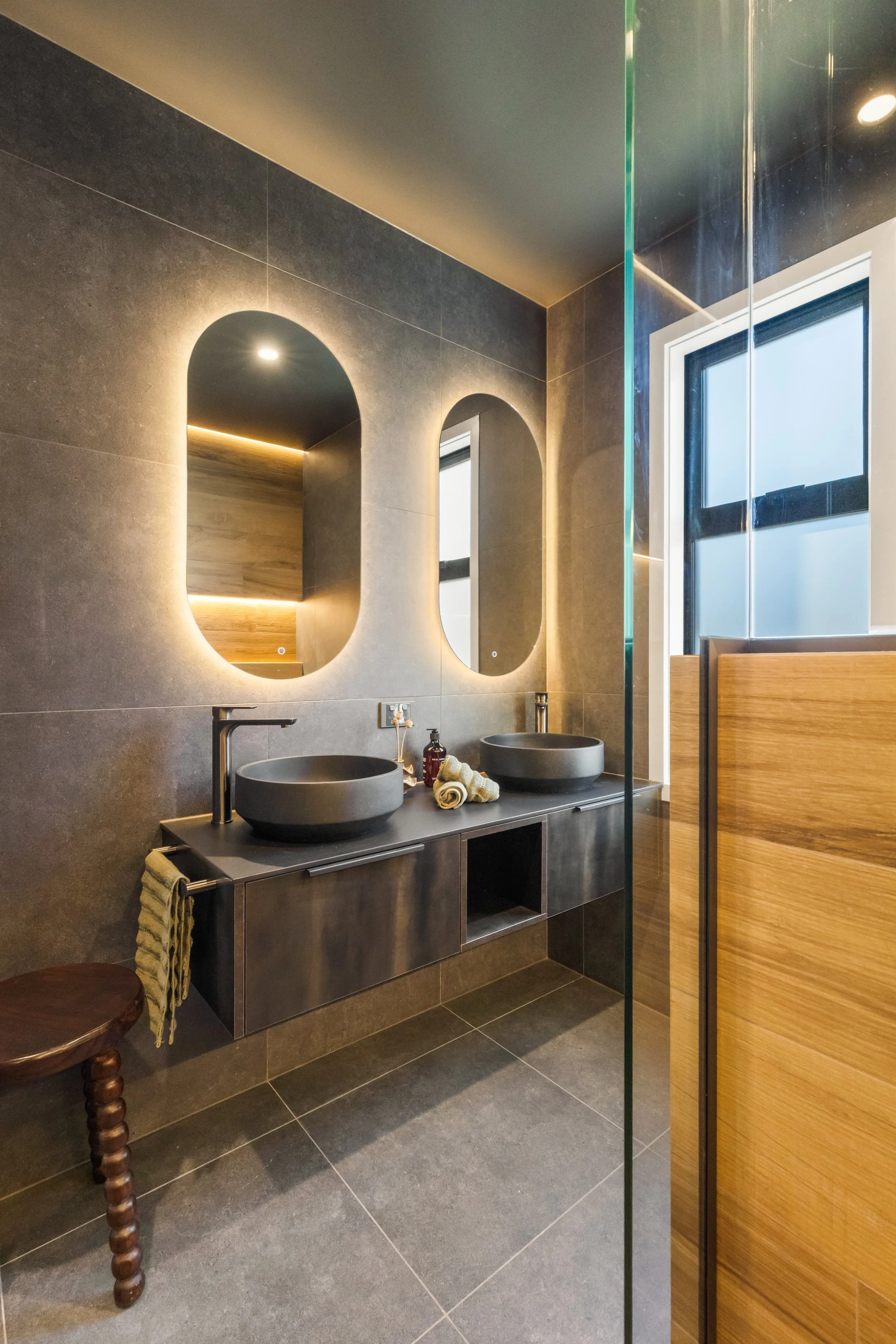 Modern bathroom with double gray vessel sinks on a gray floating vanity, large rectangular mirrors, dark gray tiled walls and floor, and a wooden privacy screen. Towel and soap on the vanity, window with black frame and cubed glass, and small wooden 