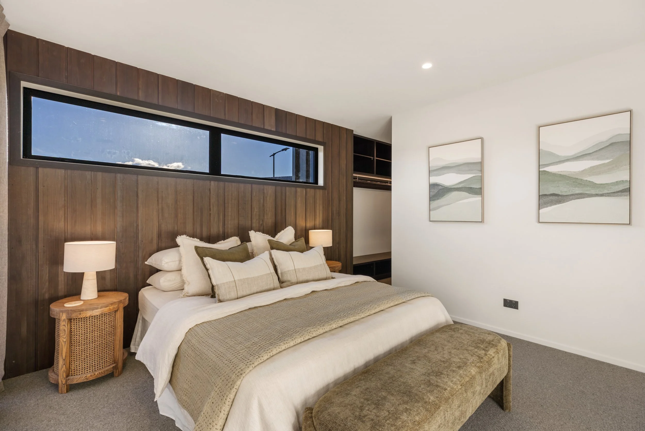 Interior of a bedroom with a bed that has multiple pillows, a wooden side table with a lamp, a dark wood-paneled wall, and framed landscape artwork on a white wall. A long horizontal window shows the sky and clouds.
