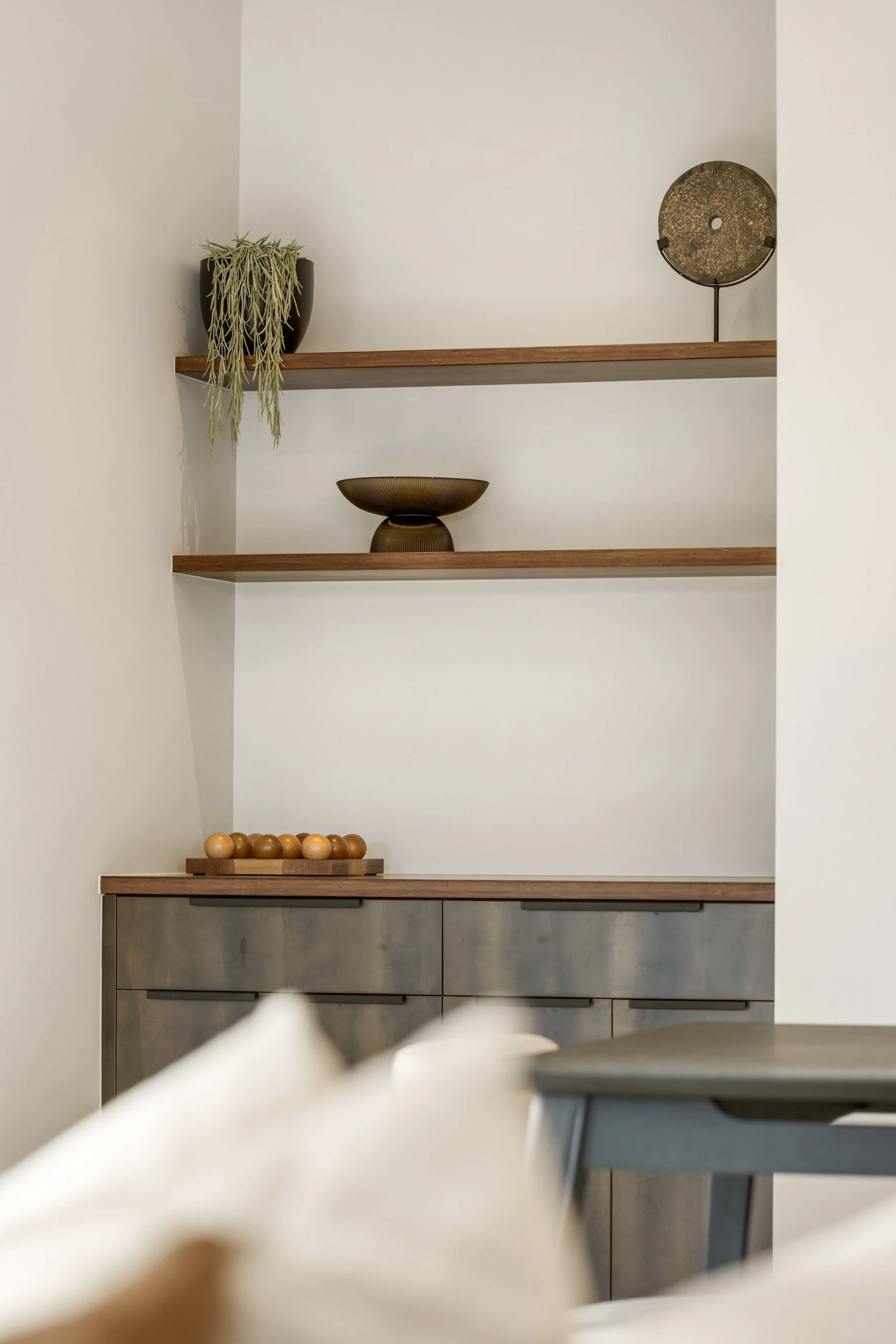 Interior view of a modern kitchen with open wooden shelves holding decorative items, including a potted plant, a round metallic bowl, and a large vintage-style clock, with cabinets below and a table or countertop in the foreground.