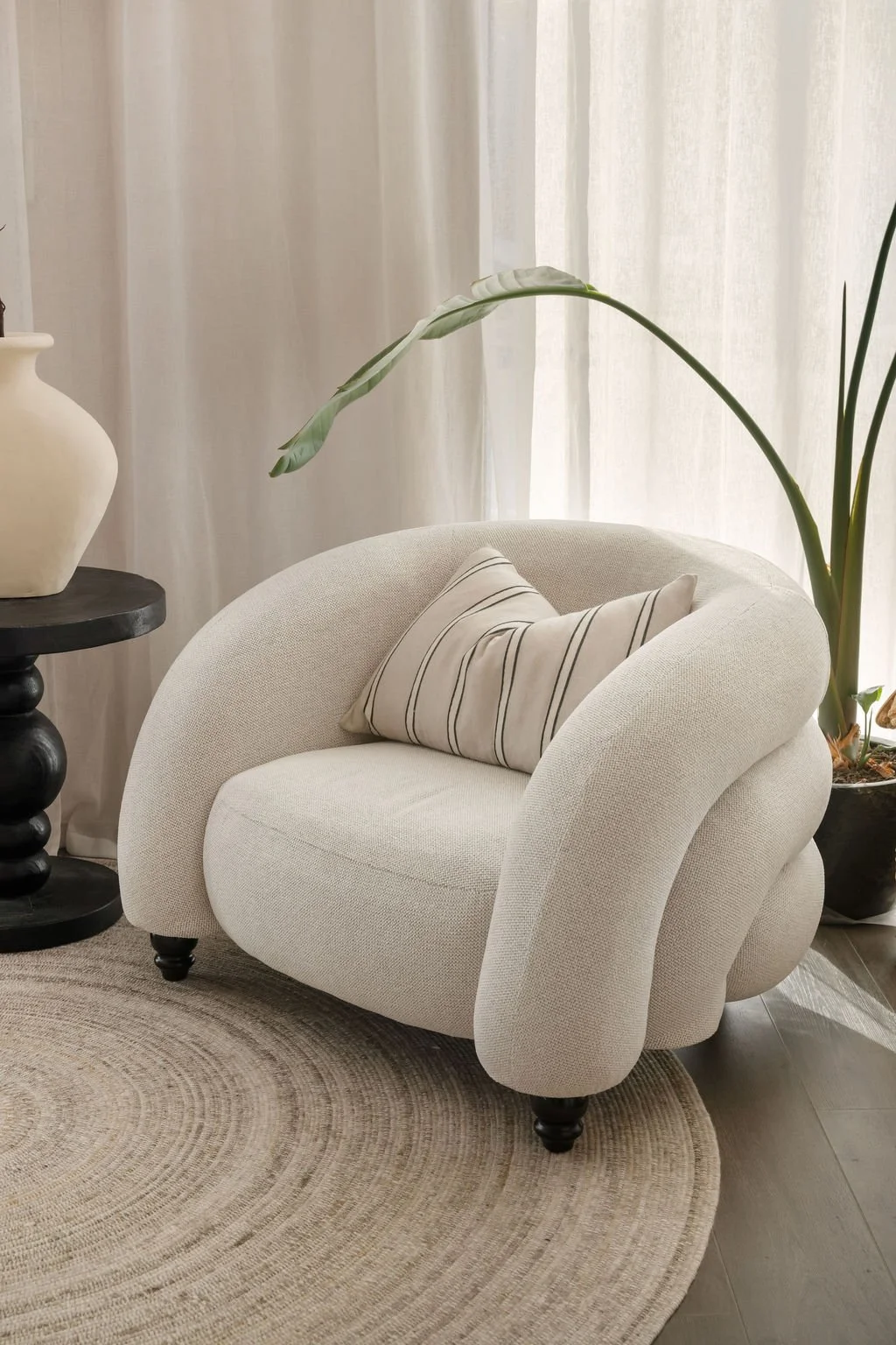 A cozy modern living room corner with a white textured armchair, a striped throw pillow, a black side table with a cream vase, and a large plant with broad leaves near sheer curtains.