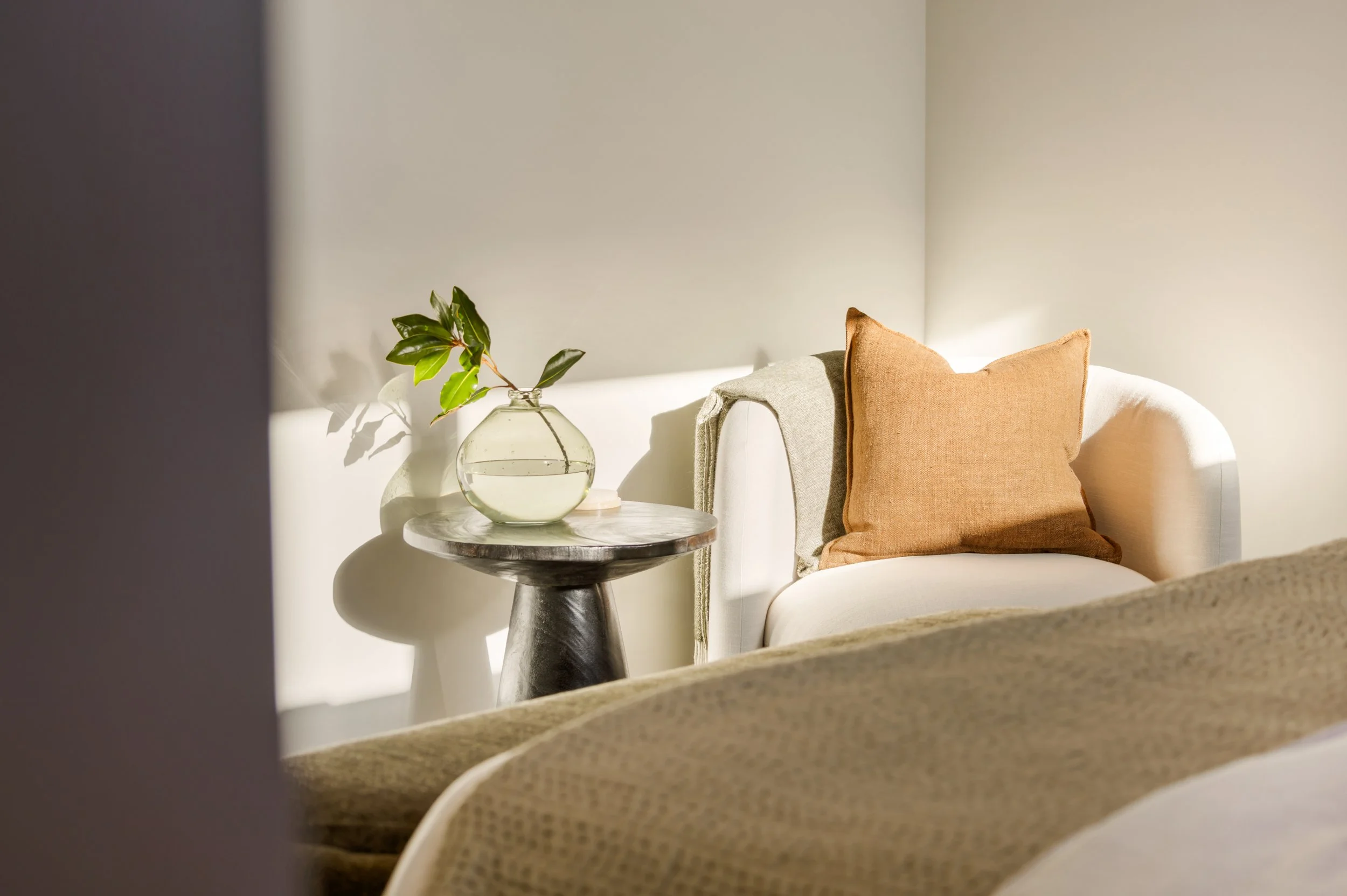 A cozy corner of a living room with a white sofa, a brown pillow, a beige blanket, a marble-top side table, a glass vase with green leaves, and sunlight casting shadows on the wall.