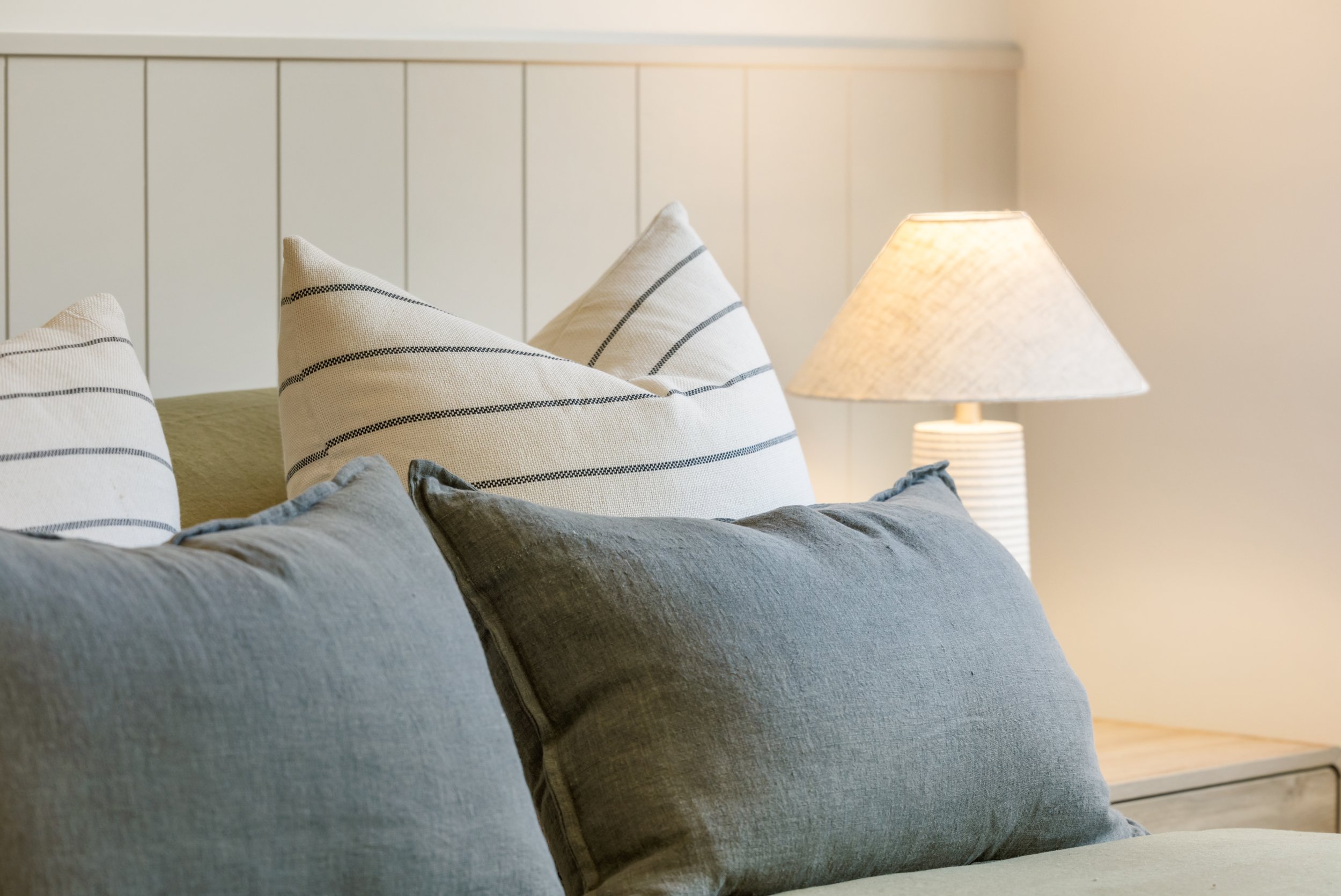 A cozy bedroom corner with a beige upholstered bed, white pillows with black striped patterns, and a bedside lamp with a textured cream lampshade.